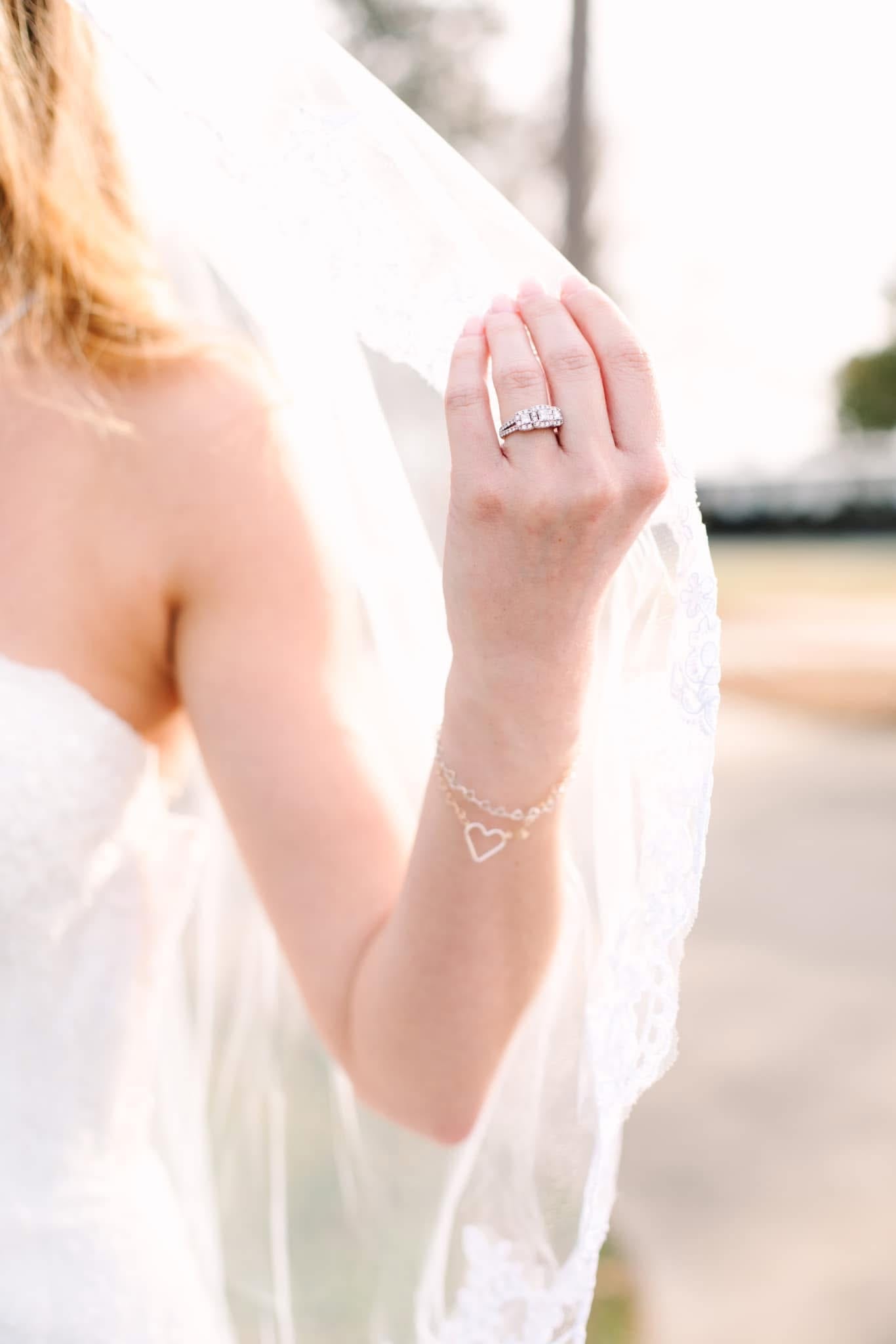 Close-up of a bride's hand holding her flowing white veil, wearing a sparkling diamond wedding ring set and a delicate heart charm bracelet in warm golden light.