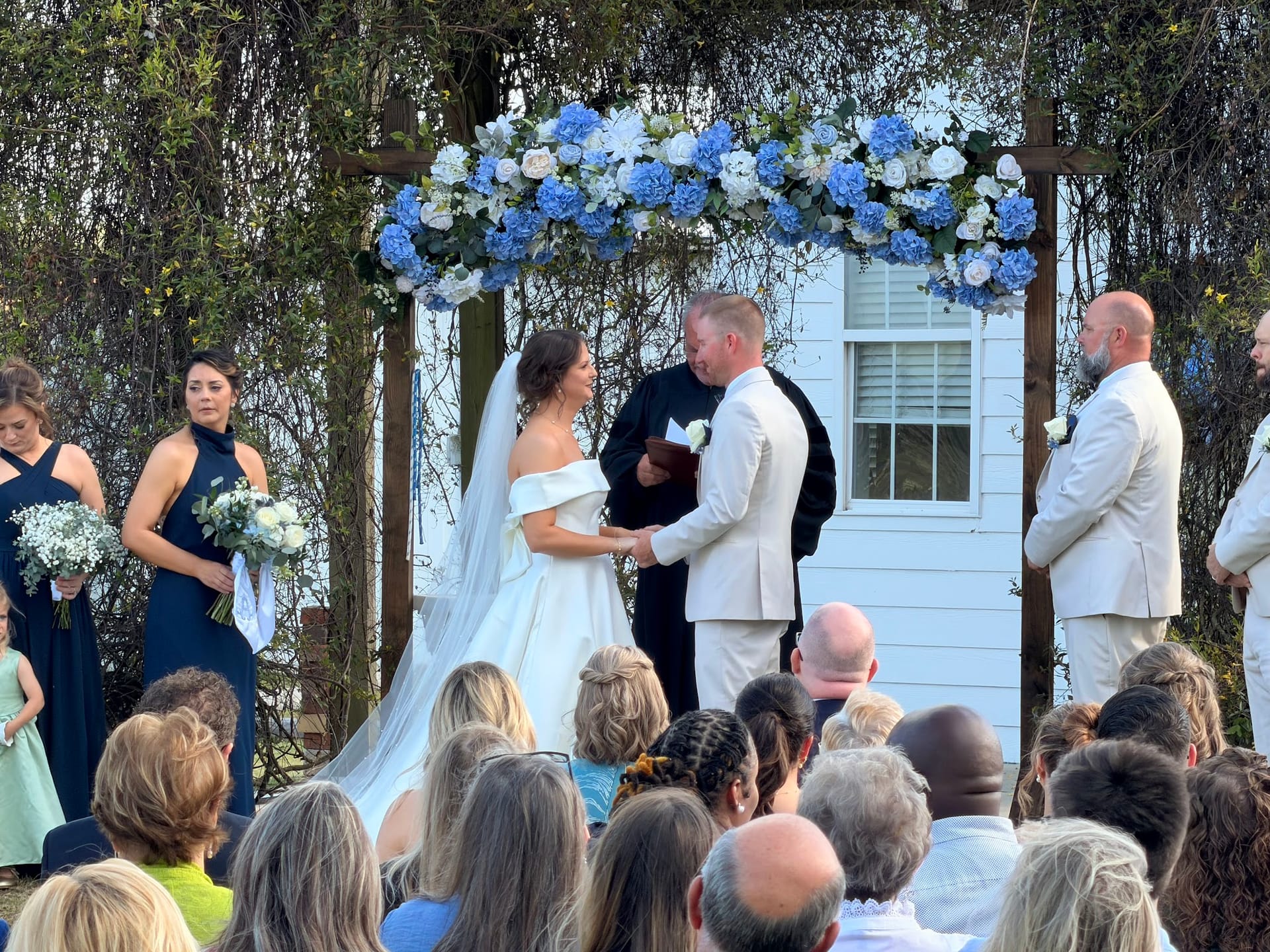 Bride and groom hold hands during their outdoor wedding ceremony beneath a rustic wooden arch adorned with blue hydrangeas and white roses at Scarborough House historic estate.