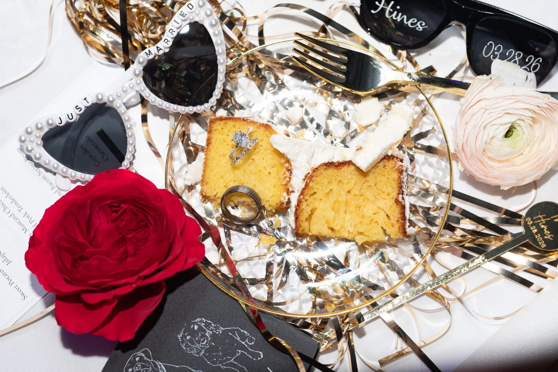 Flat-lay wedding detail shot featuring slices of white cake with diamond and band rings resting on top, pearl heart-shaped 'Just Married' sunglasses, personalized 'Hines 03.28.26' sunglasses, a red rose, a blush ranunculus, a gold fork, and gold confetti streamers arranged on a glass plate.
