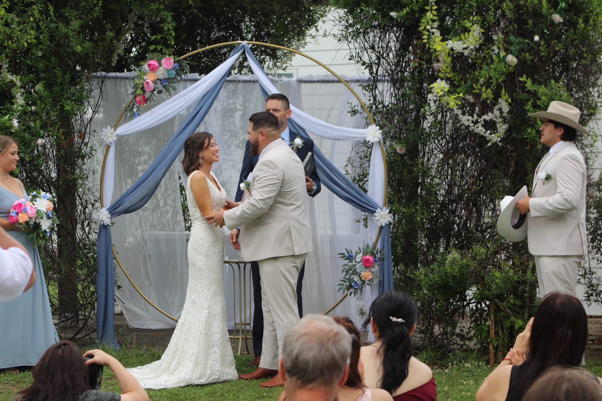 Smiling bride and groom exchange vows beneath a gold hoop arch draped in dusty blue fabric and florals on the ceremony lawn at Scarborough House.