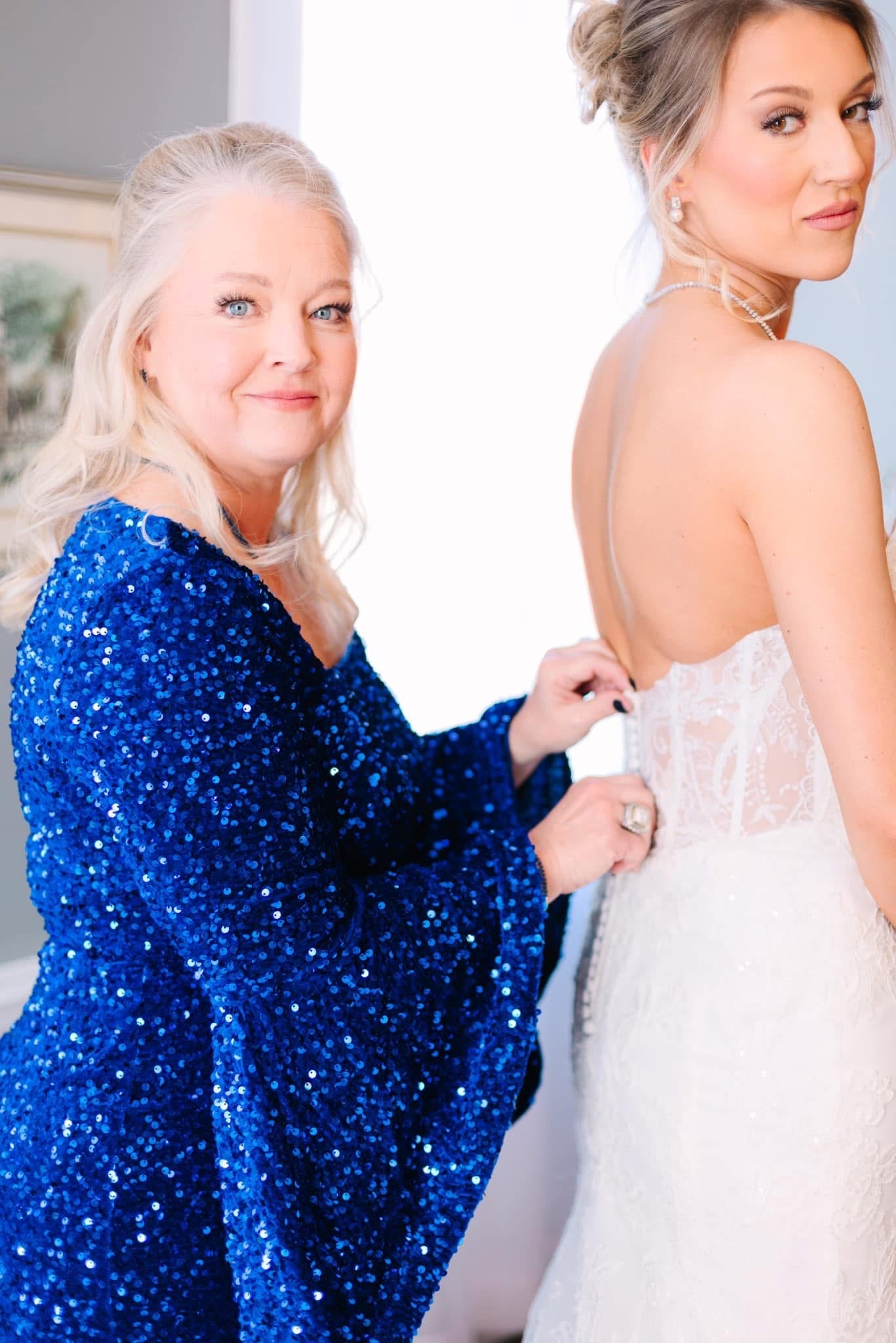 A woman in a sparkling royal blue sequin gown lovingly buttons the back of a bride's lace wedding dress during bridal preparations at Scarborough House.