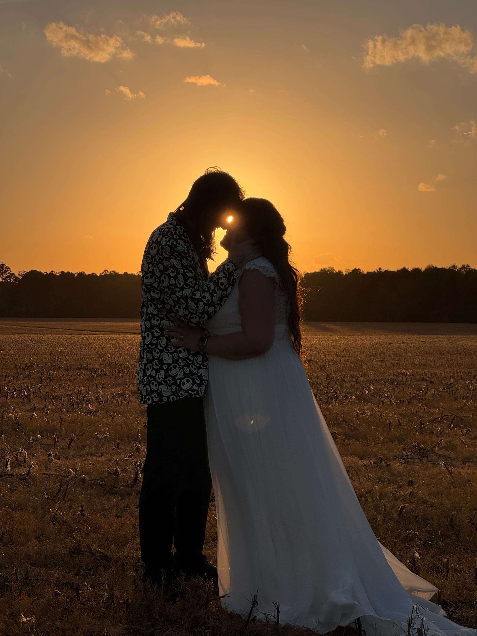 Silhouette of bride in flowing white gown and groom in patterned jacket touching foreheads against a glowing golden sunset over open farmland fields at Scarborough House.