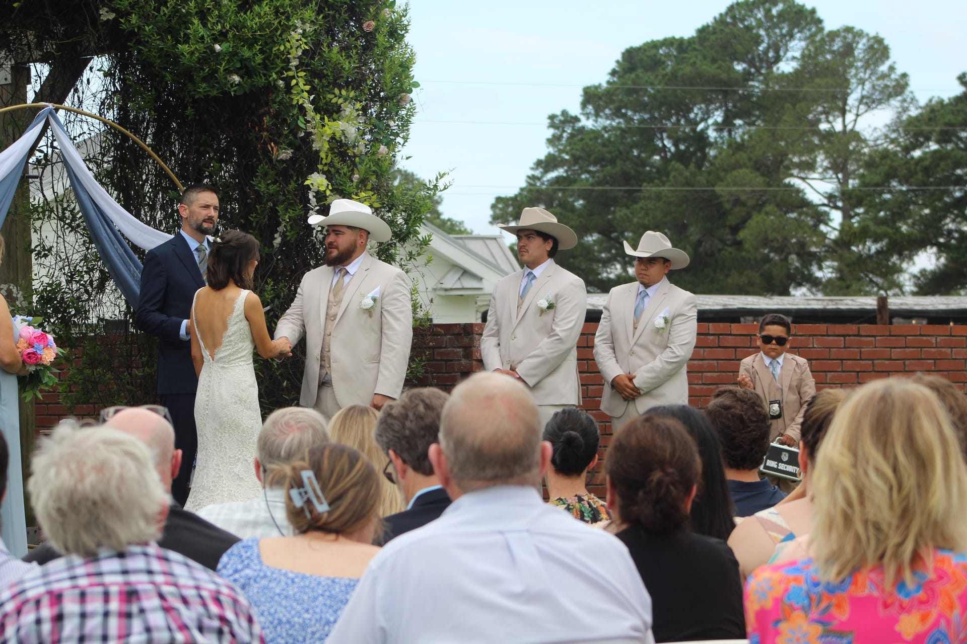Outdoor wedding ceremony at Scarborough House with three groomsmen in tan suits and cowboy hats standing at the altar as the bride and groom exchange vows before seated guests.