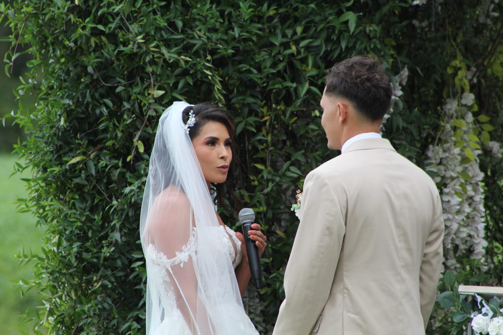 Bride in a lace gown and veil speaks heartfelt vows into a microphone while facing her groom before a lush green floral arch at an outdoor wedding ceremony.