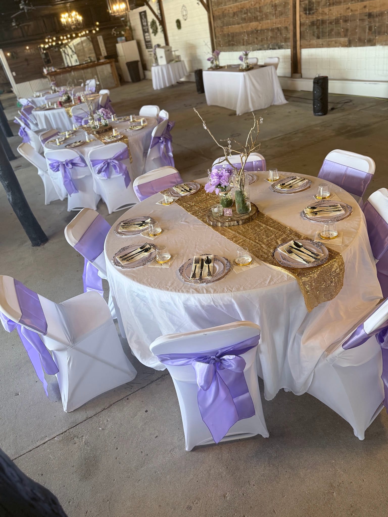 Elegantly set round reception tables with white linens, gold sequin runners, lavender chair sashes, gold cutlery, and floral centerpieces inside a rustic barn wedding venue.