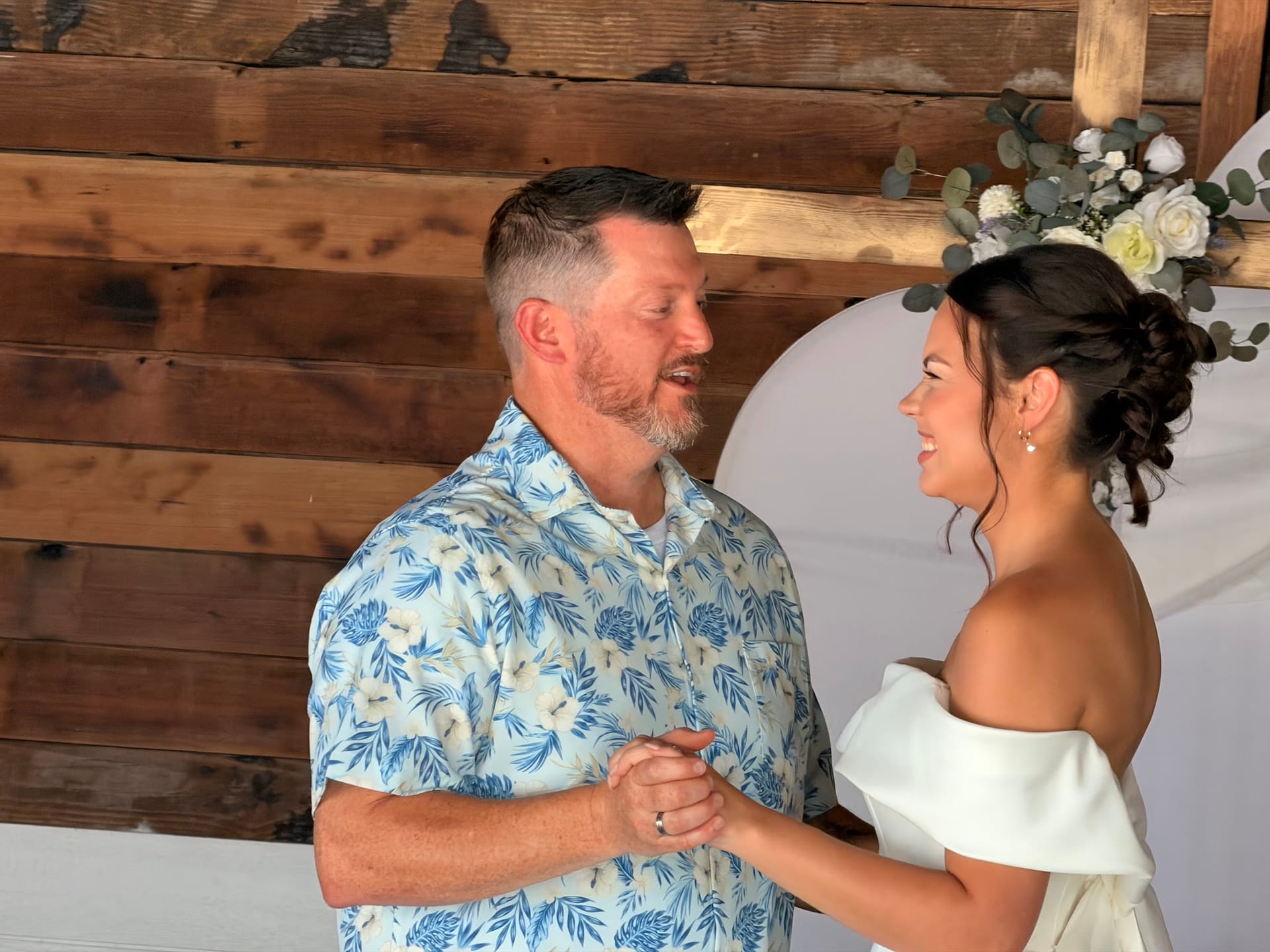 Bride in an off-shoulder white gown and groom in a blue floral shirt share a joyful first dance beneath a floral arch against a rustic wood-plank backdrop at Scarborough House.