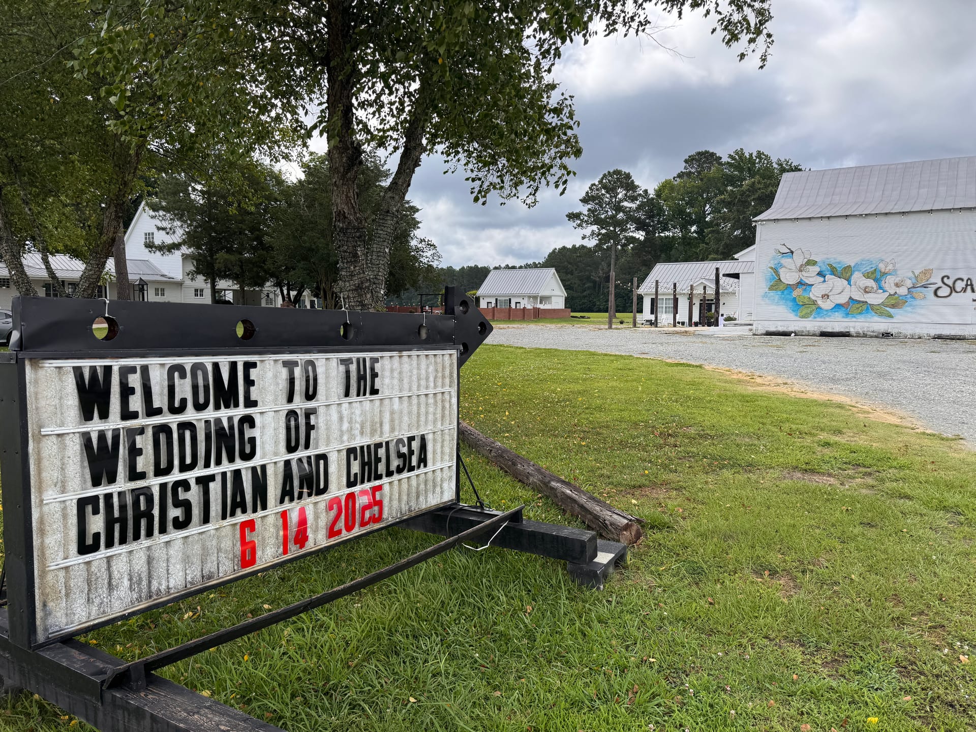 Marquee sign on the grounds of Scarborough House welcoming guests to the wedding of Christian and Chelsea on June 14 2025, with a white barn featuring a magnolia mural in the background.