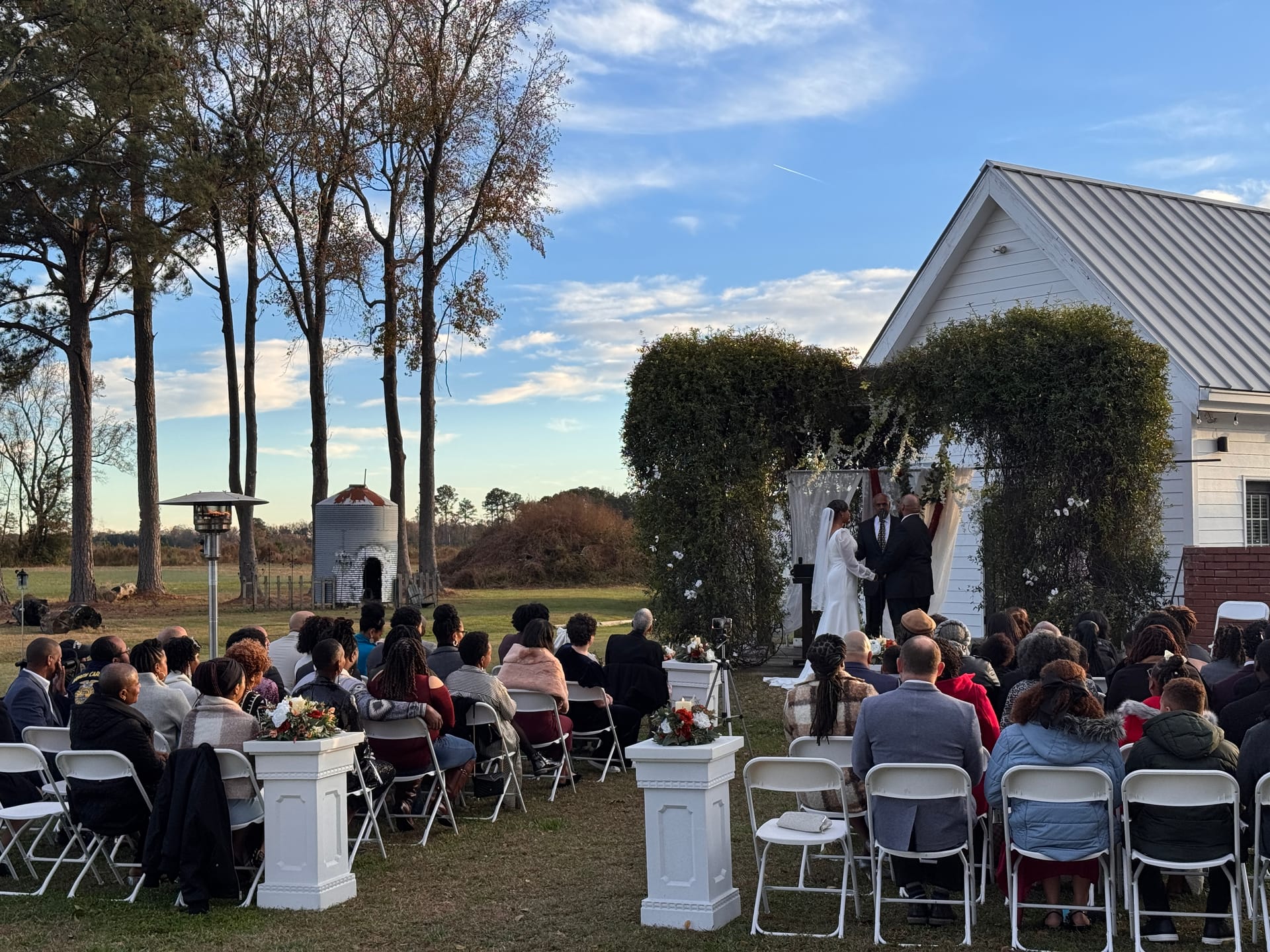 Guests seated in white folding chairs watch a couple exchange vows beneath a lush green floral arch on the ceremony lawn at Scarborough House estate with a white chapel and open farmland fields in the background.