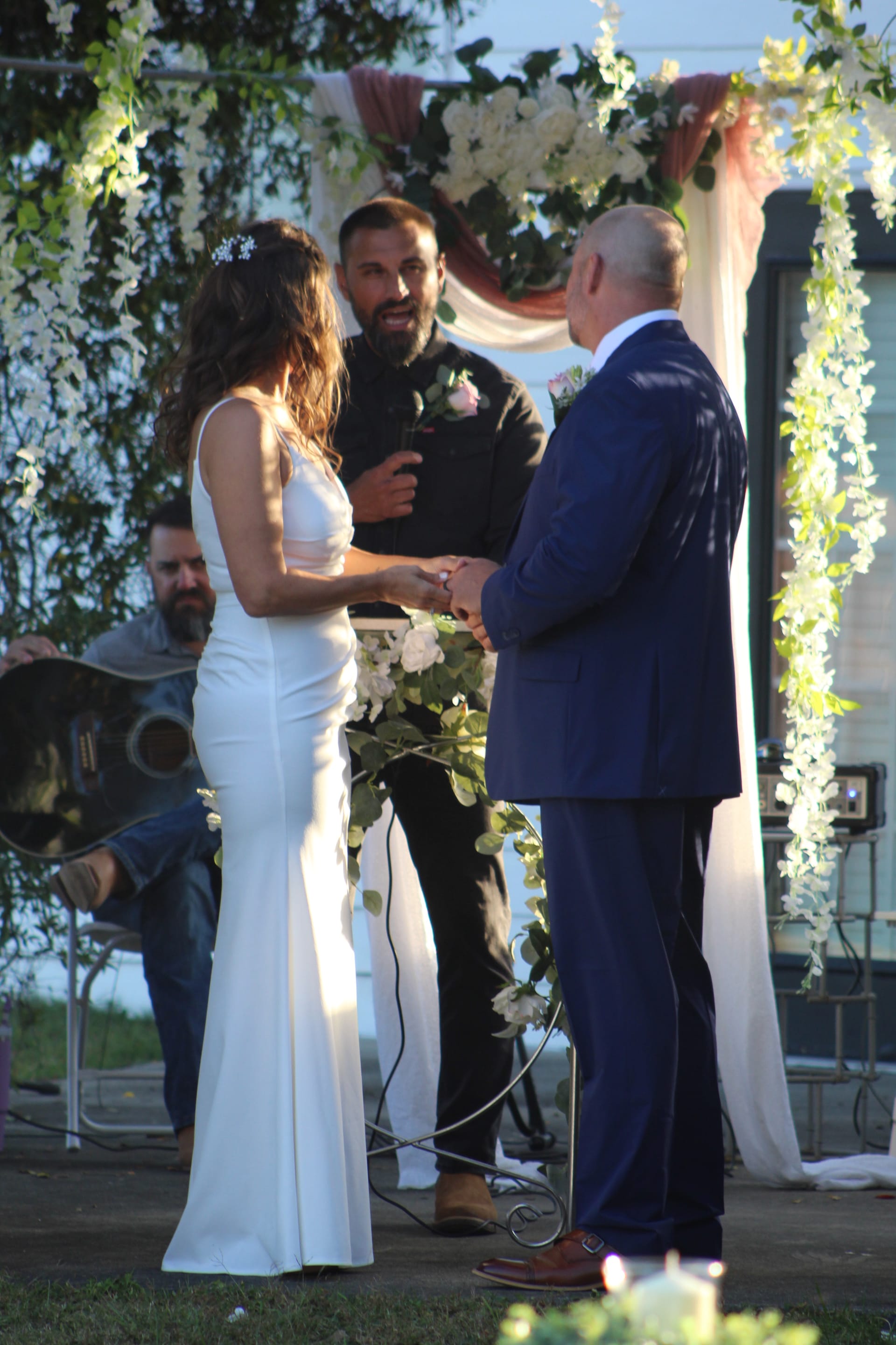 Bride in a fitted white gown and groom in a navy suit hold hands during their outdoor wedding ceremony beneath a lush floral arch adorned with white blooms and draped fabric at Scarborough House.