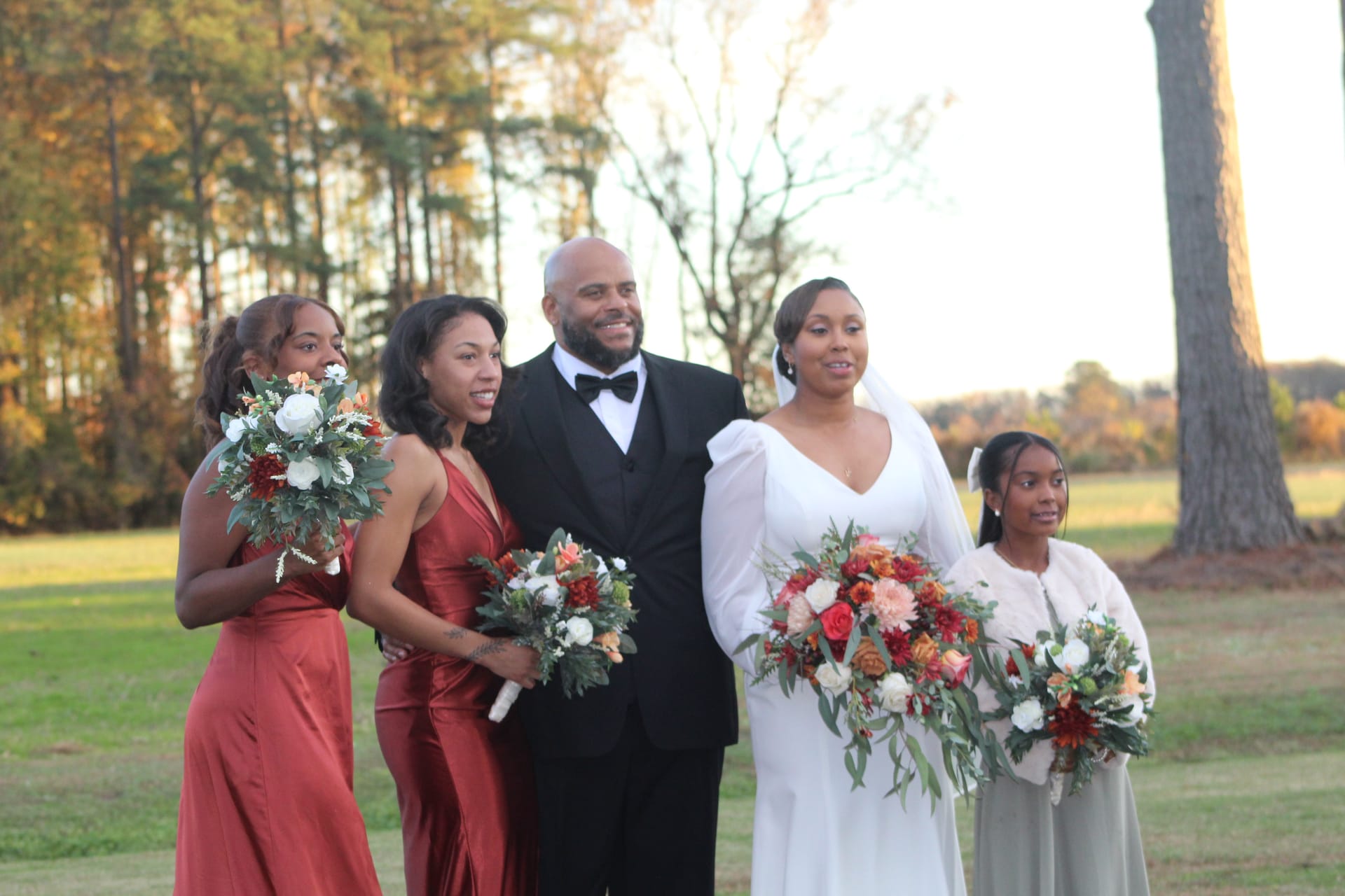 Bride in a white long-sleeve gown holds a cascading floral bouquet alongside her groom in a black tuxedo and two bridesmaids in rust-red gowns and a junior bridesmaid on an autumn ceremony lawn at Scarborough House.