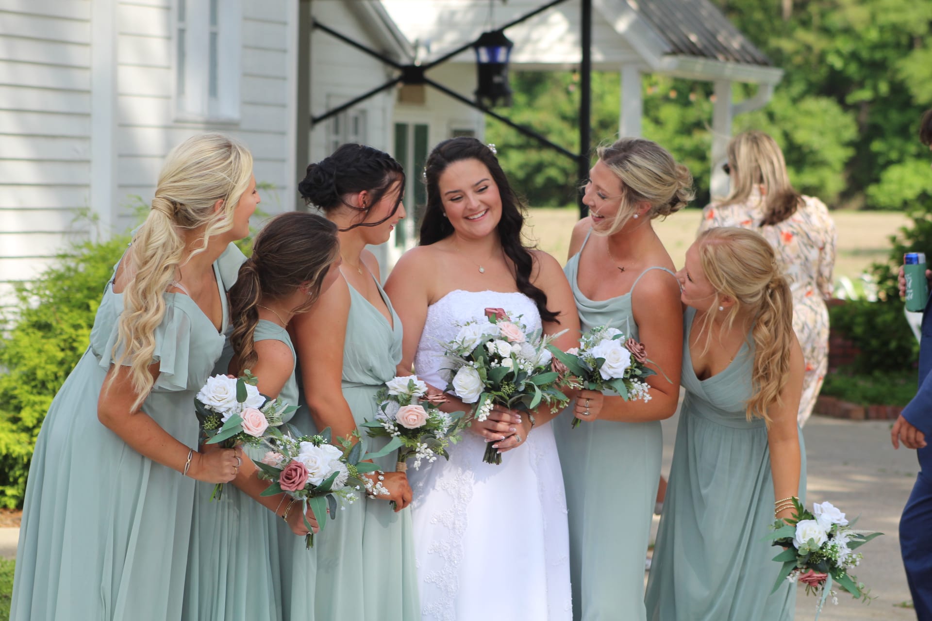 Bride in white lace gown laughs with four bridesmaids in sage green dresses holding floral bouquets outside the historic Scarborough House estate.