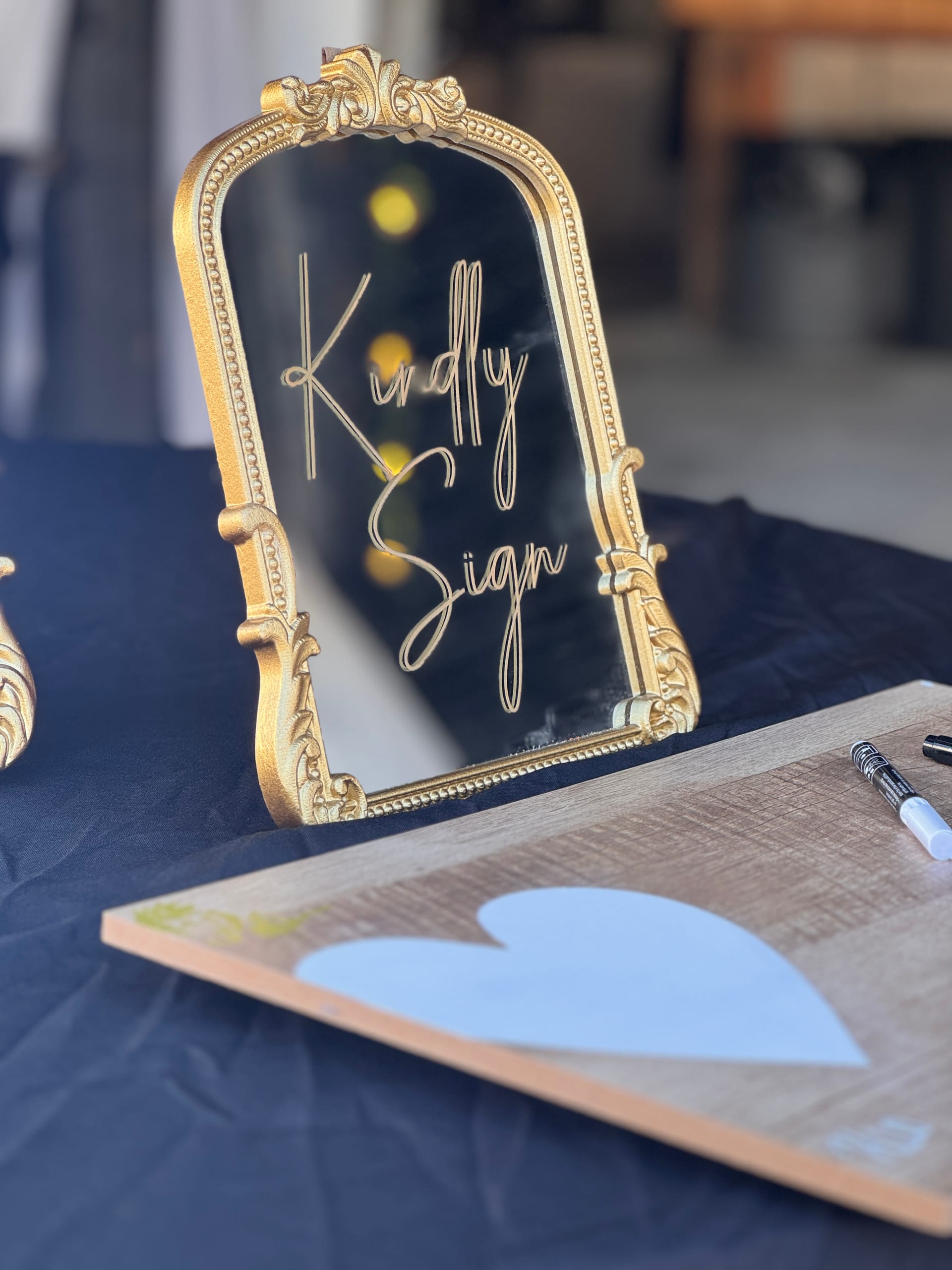 An ornate gold-framed mirror sign reading Kindly Sign displayed on a navy linen table beside a rustic wooden guest book and calligraphy markers at an elegant wedding reception.