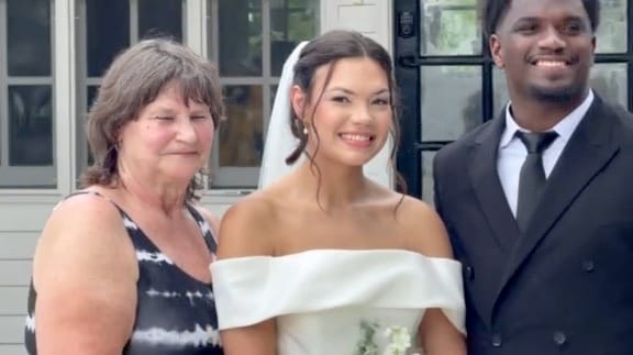 Bride in an off-shoulder white gown and veil smiles with an older woman and a man in a dark suit in front of the historic Scarborough House estate.