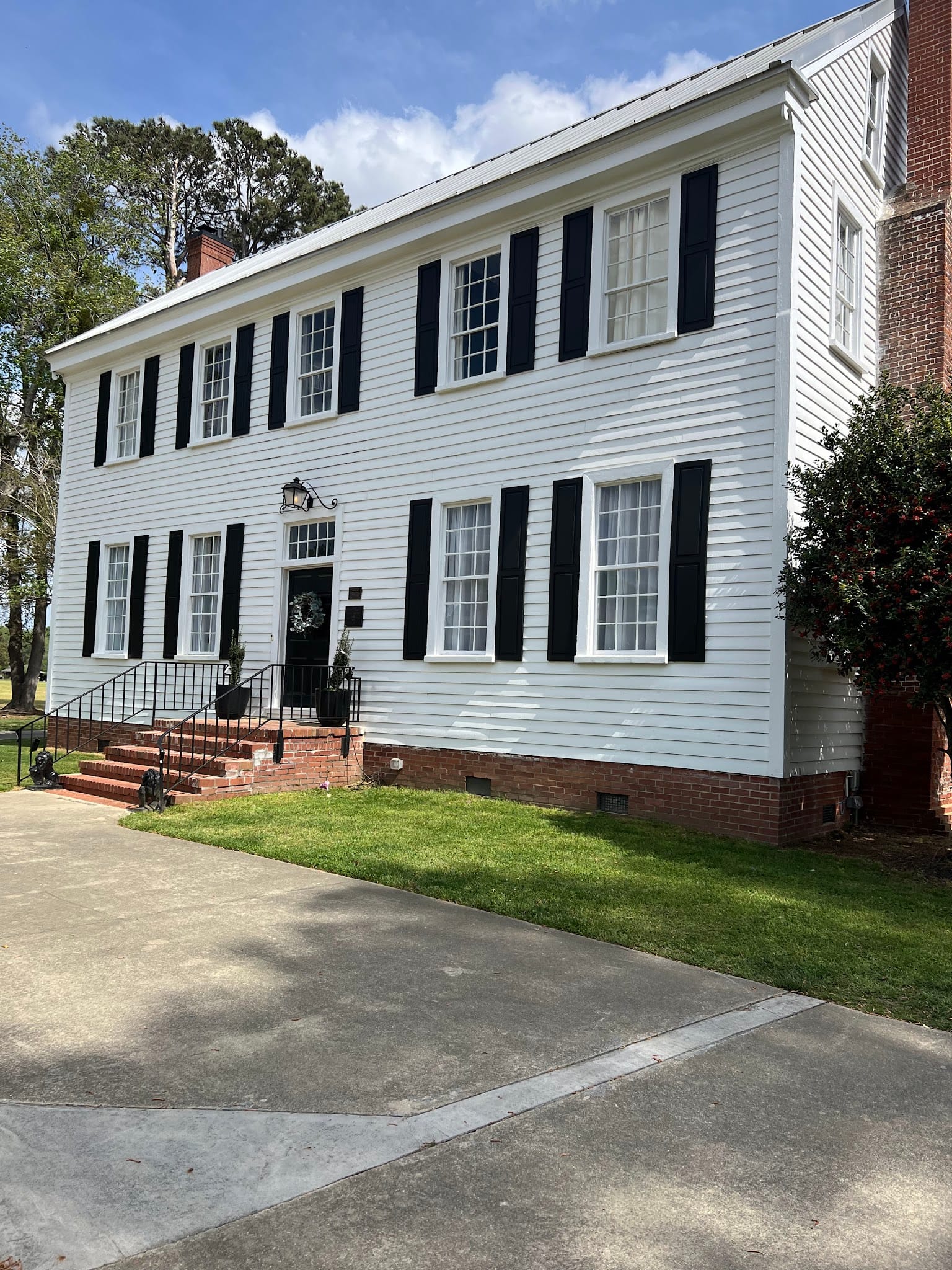 Front exterior of the historic 1821 Scarborough House wedding venue featuring white clapboard siding, black shutters, brick front steps with iron railings, and a welcoming wreath on the front door under a bright blue sky.