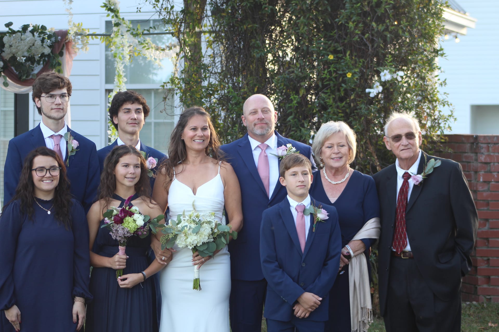 Bride in white gown and groom in navy suit pose with their blended family of seven on the ceremony lawn at Scarborough House historic wedding estate.