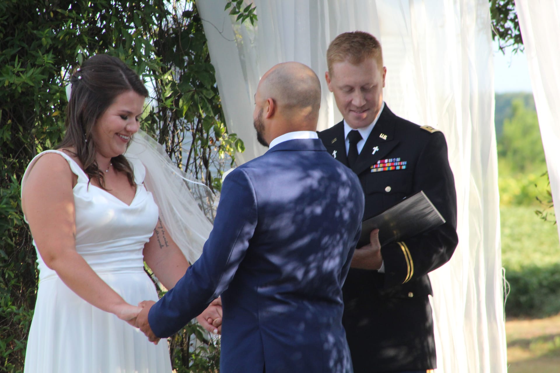 Smiling bride in white gown and veil holds hands with her groom during an outdoor wedding ceremony officiated by a military chaplain in dress uniform beneath a white draped arch.