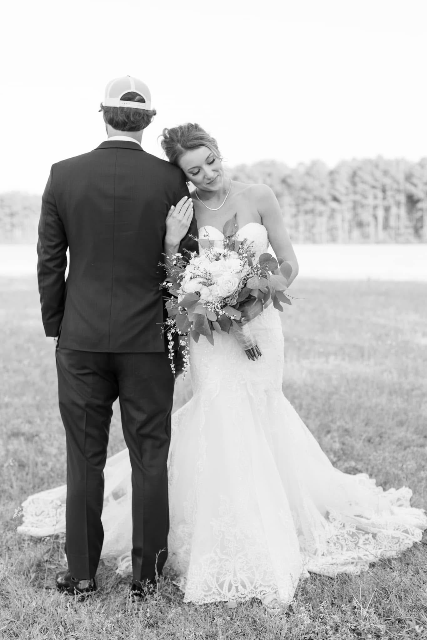 Bride in a lace mermaid gown leans affectionately against her groom in a dark suit on the open farmland fields of Scarborough House, holding a full floral bouquet while her cathedral train spreads across the grass.