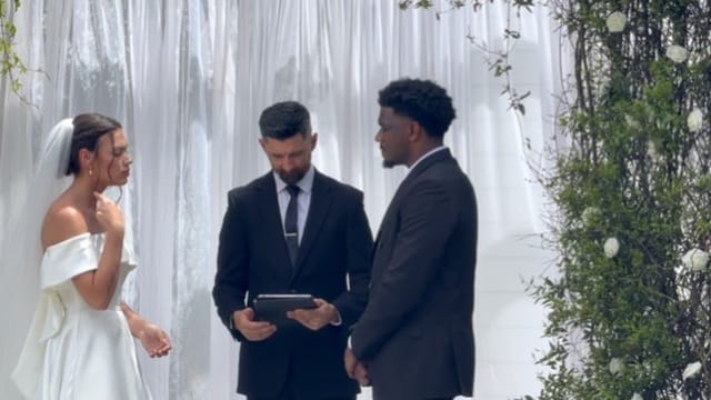Bride in off-shoulder white gown and groom in dark suit exchange vows before an officiant beneath a draped white backdrop with white floral accents at an outdoor wedding ceremony.