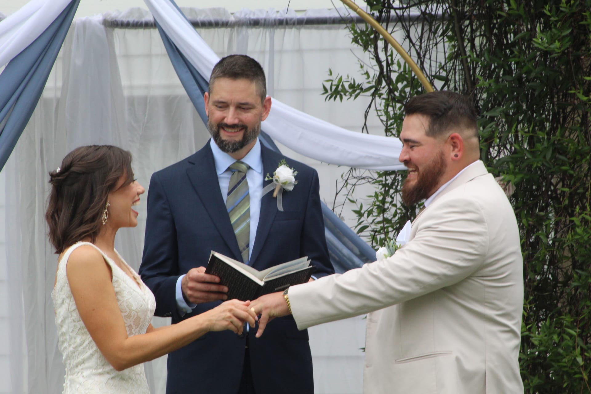 Bride and groom exchange rings and share a laugh during their outdoor wedding ceremony beneath a draped arch at Scarborough House.