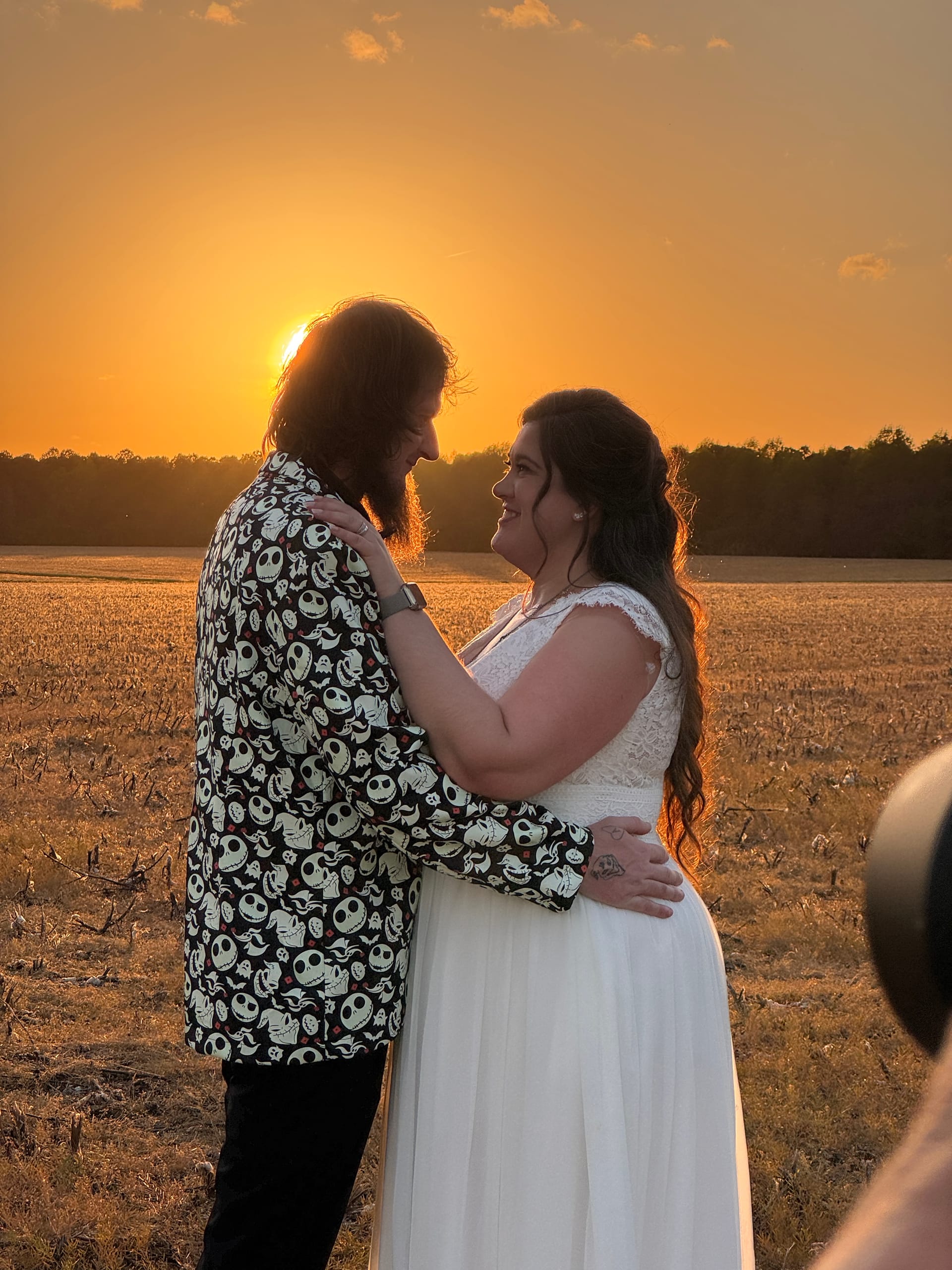 Bride in white lace gown and groom in a Nightmare Before Christmas blazer share a tender moment at golden hour in a farmland field at Scarborough House.