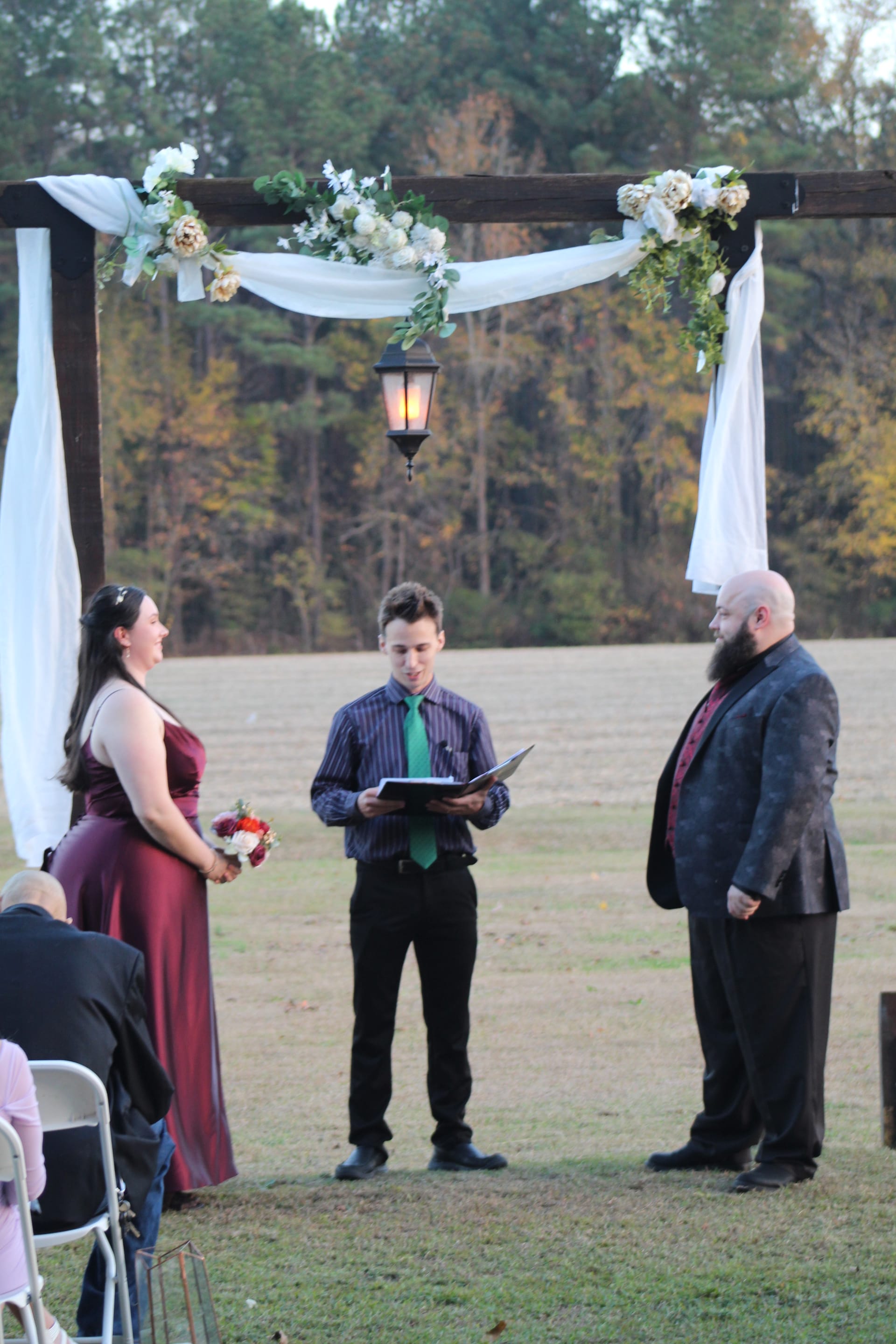 A bride in a burgundy gown holding a bouquet stands with a bearded groom and officiant beneath a rustic wooden arch draped in white fabric and white florals on the ceremony lawn at Scarborough House.
