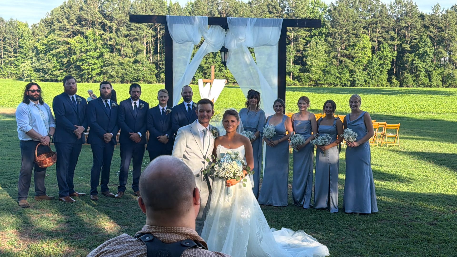 Bride and groom stand together under a white-draped wooden arch on a sunlit lawn surrounded by groomsmen in navy suits and bridesmaids in dusty blue gowns holding white floral bouquets with a pine tree forest in the background.