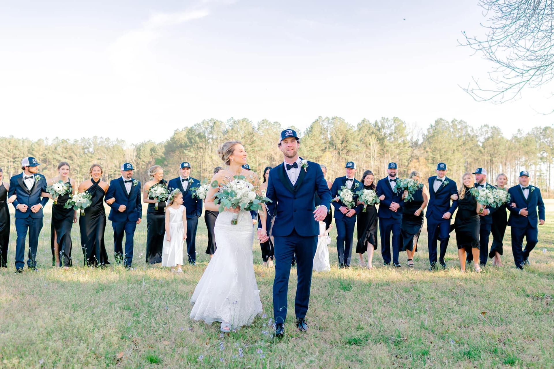 Bride and groom walk hand in hand leading their large wedding party across a sunlit open field at Scarborough House historic estate venue.