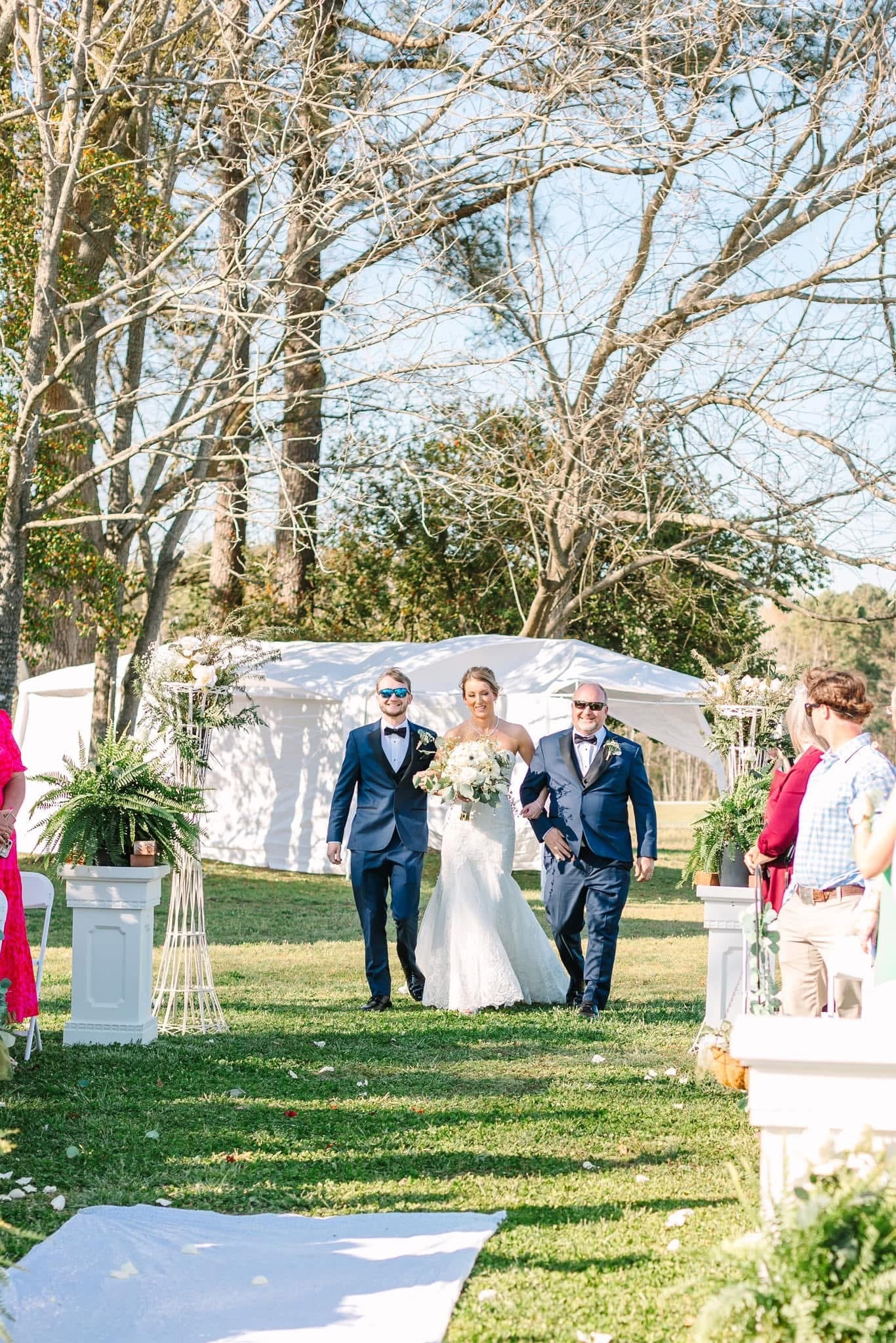 Smiling bride in a lace gown holding a white bouquet walks down the outdoor aisle flanked by two men in navy suits at Scarborough House ceremony lawn.