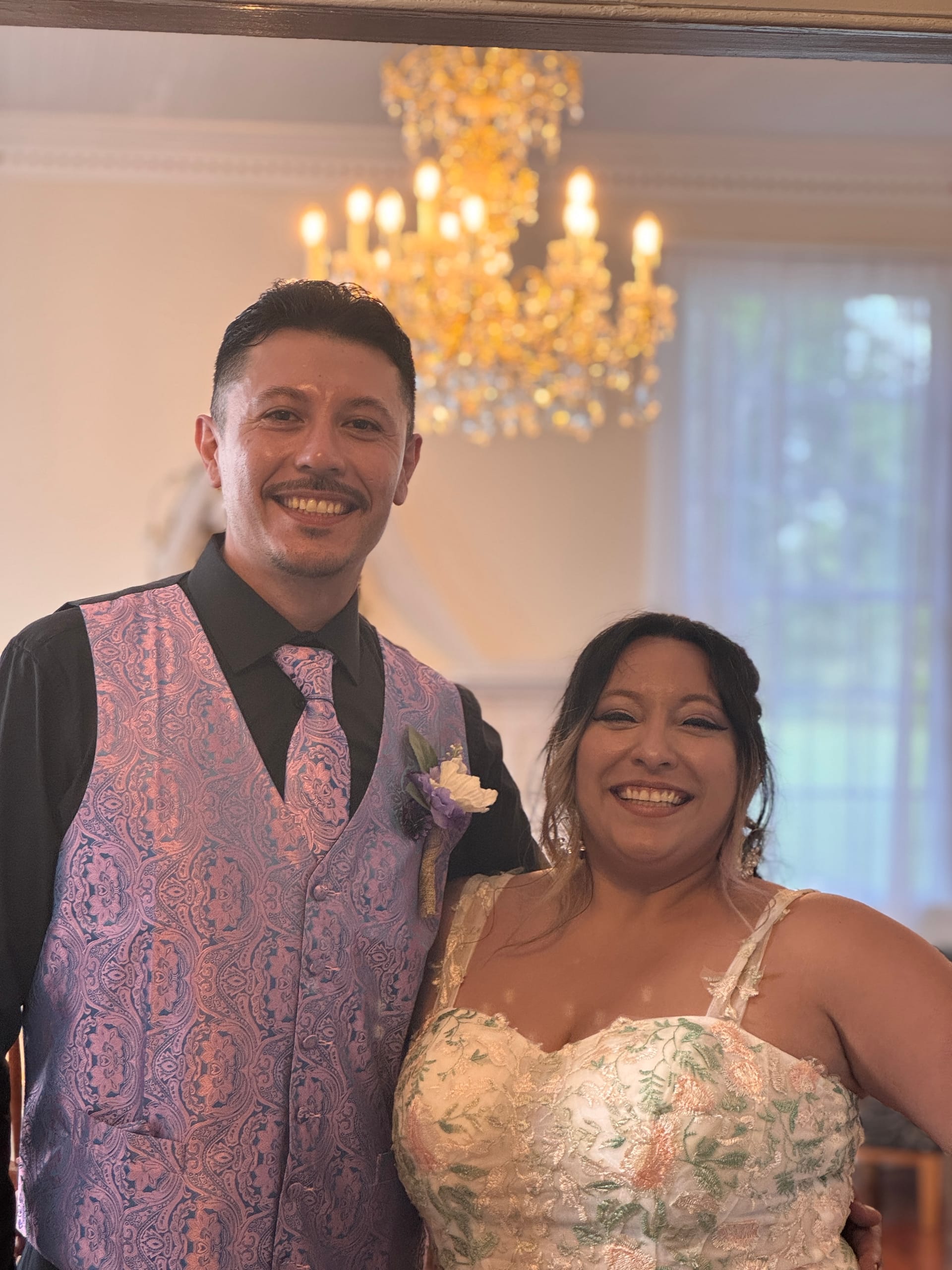Smiling bride in a floral embroidered gown and groom in a paisley vest and matching tie pose together beneath a glowing chandelier inside the grand hall at Scarborough House.