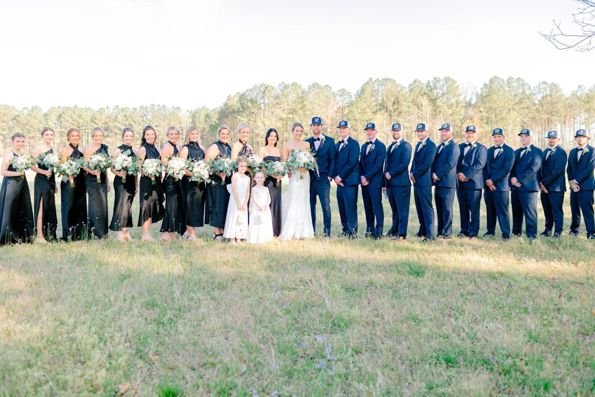 Large wedding party in black bridesmaid dresses and navy suits with baseball caps lines up across a sunlit open field at Scarborough House with tall pines in the background.