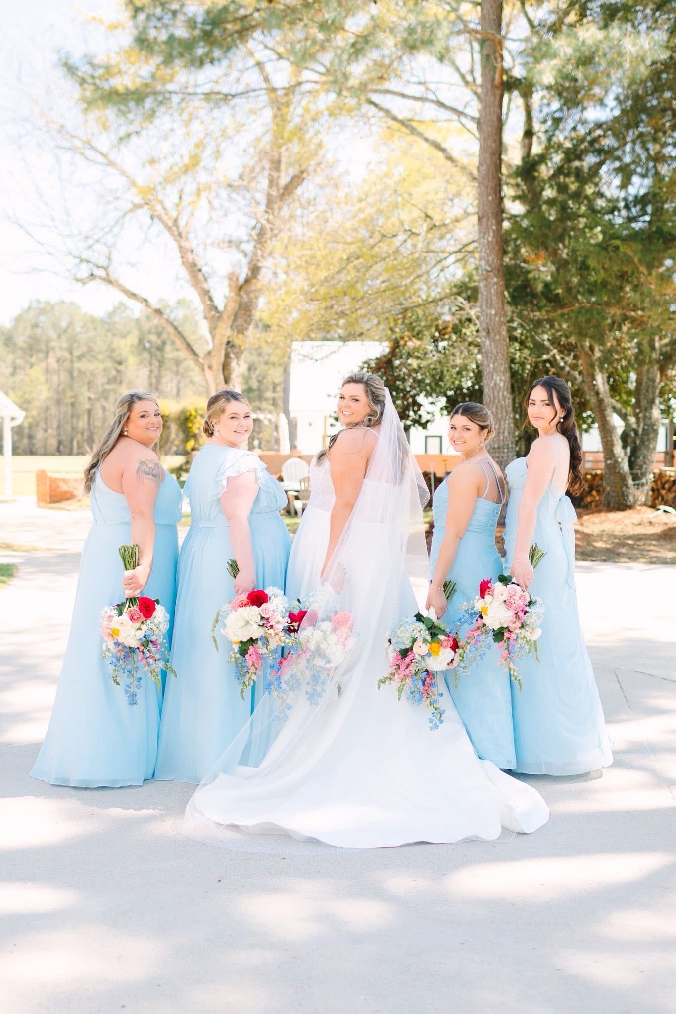 Bride in a white gown and veil looks back over her shoulder surrounded by four bridesmaids in light blue dresses holding colorful floral bouquets on the grounds of Scarborough House wedding venue.