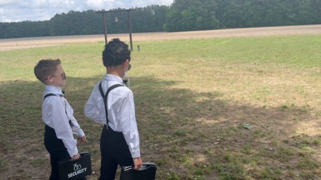 Two young boys dressed in white shirts suspenders and sunglasses carrying security briefcases stand facing the open farmland fields at Scarborough House wedding venue.
