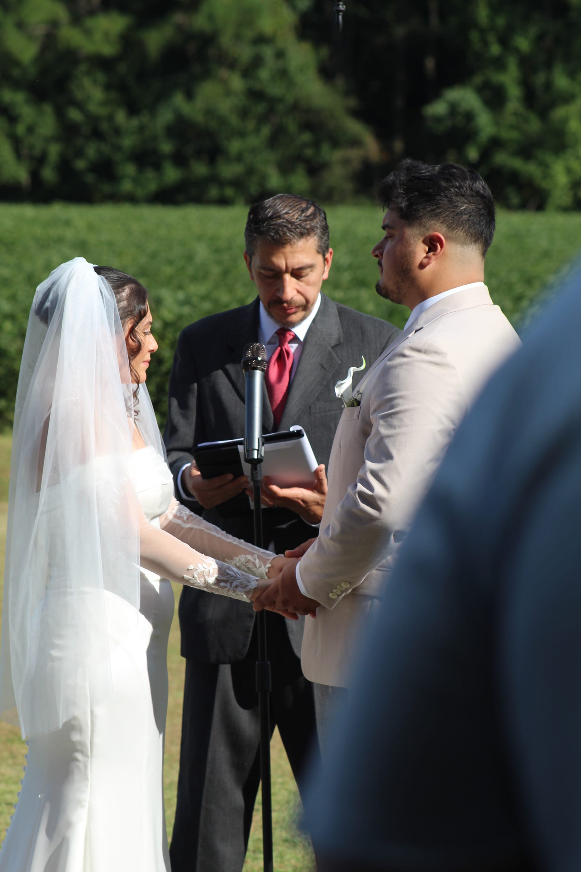 Bride in white lace gown and veil holds hands with groom in cream suit as officiant reads from a tablet during an outdoor wedding ceremony at Scarborough House.