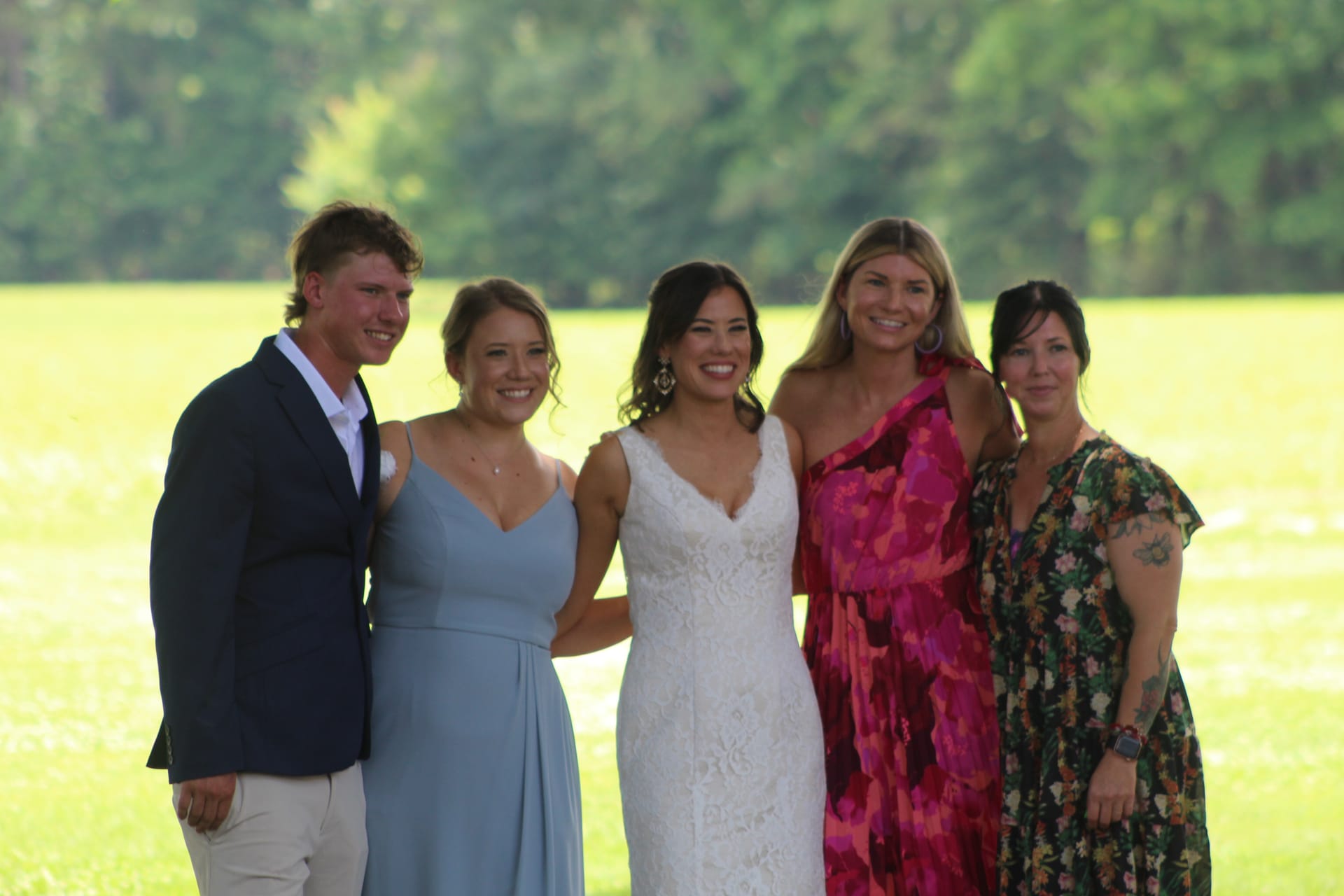 Bride in white lace gown smiling with four guests including a bridesmaid in blue and a woman in pink floral dress on a sunlit green lawn at Scarborough House.