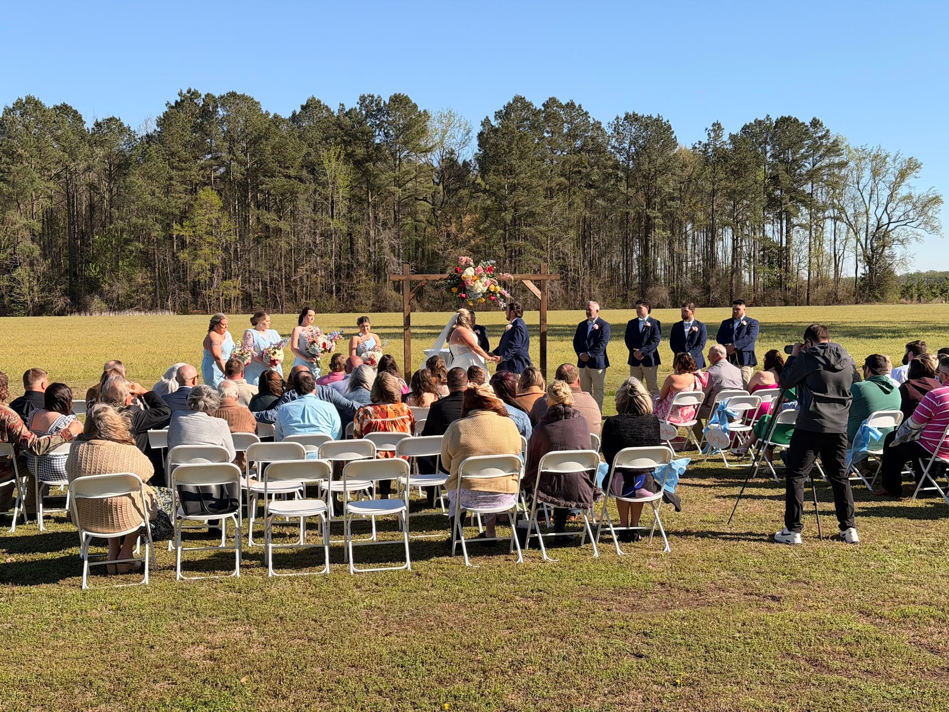 Outdoor wedding ceremony on a sunny day at a scenic farmland estate with guests seated in white folding chairs facing the couple and wedding party at a rustic wooden arch adorned with colorful flowers.