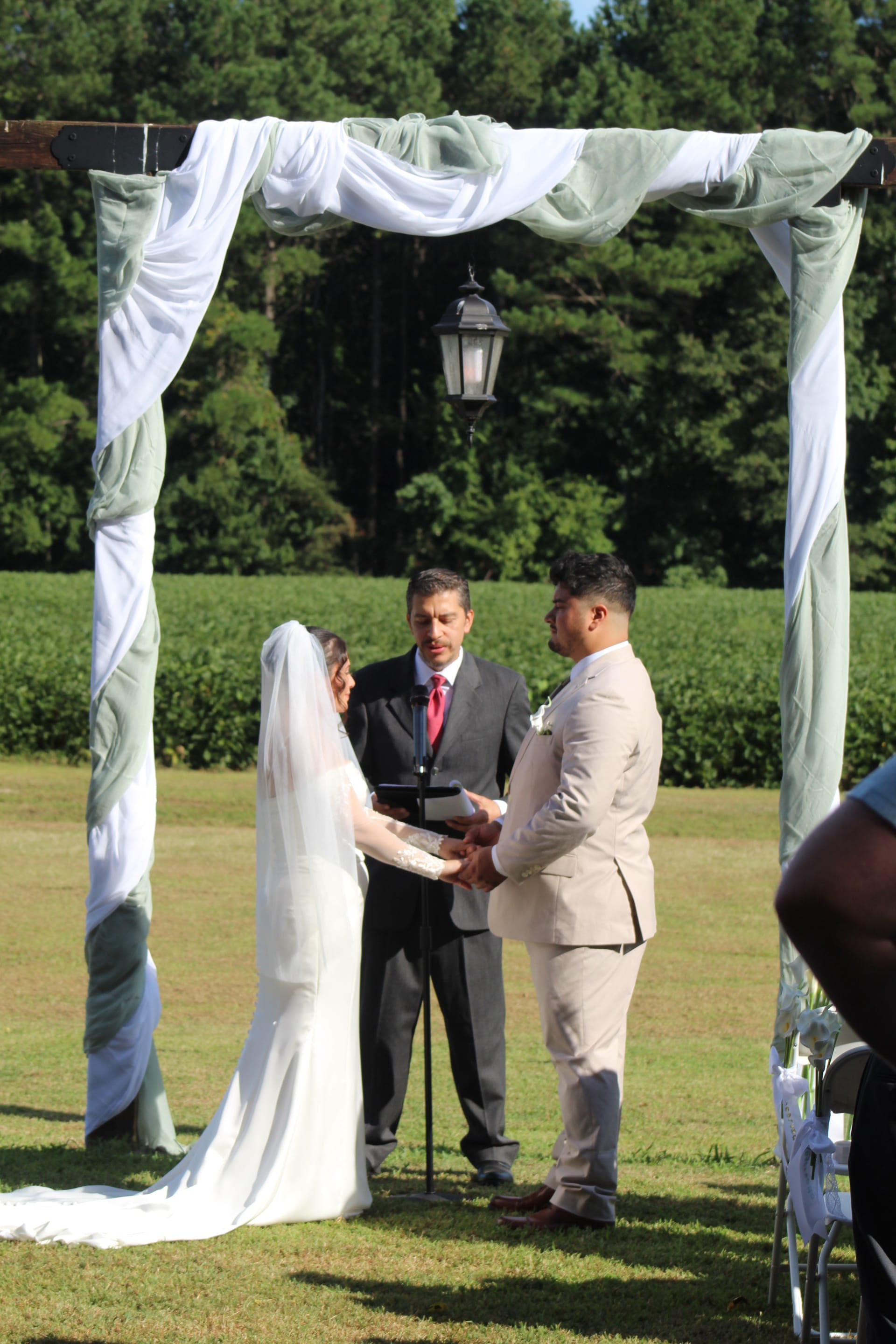 Bride and groom exchange vows beneath a draped wooden arch adorned with white and sage green fabric on the ceremony lawn at Scarborough House, with lush farmland fields and pine trees in the background.
