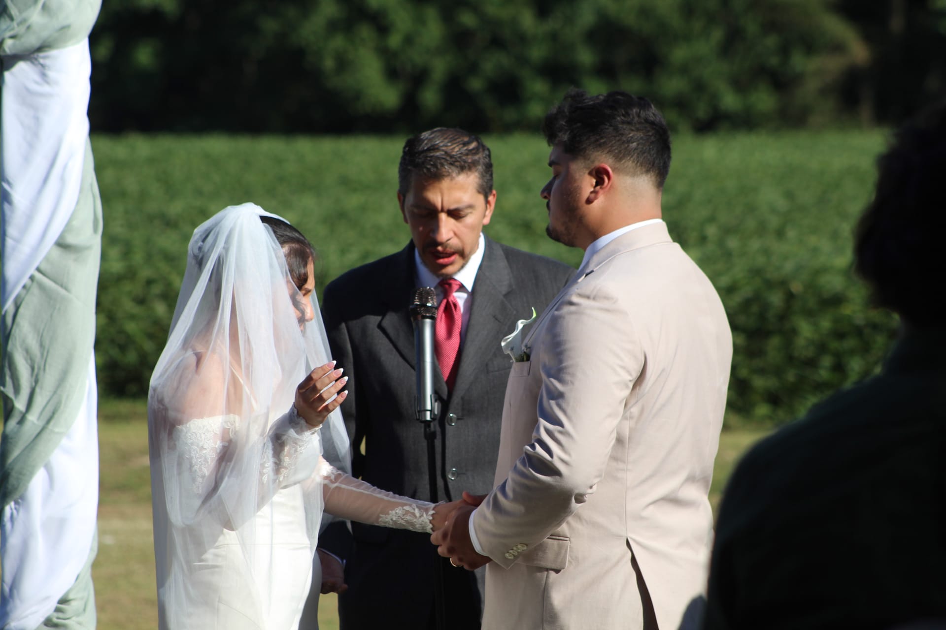 Bride in lace gown and veil holds hands with groom in cream suit as officiant speaks during an outdoor wedding ceremony on the estate lawn surrounded by lush green fields.