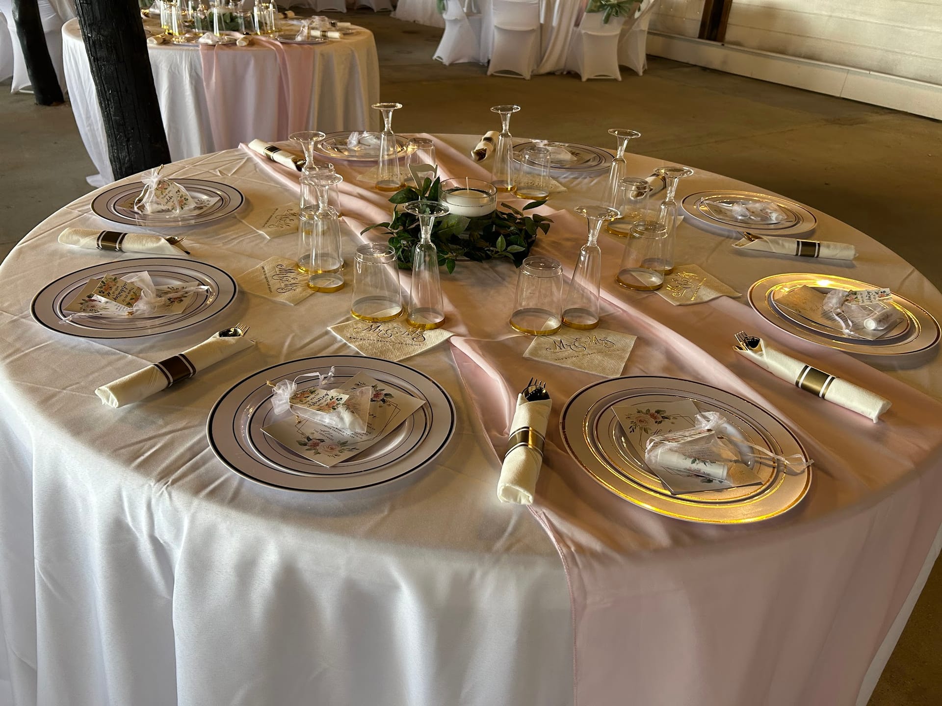 Elegantly set round reception table with gold-rimmed charger plates, rolled linen napkins, crystal stemware, and a greenery candle centerpiece on a blush satin runner inside Scarborough House.