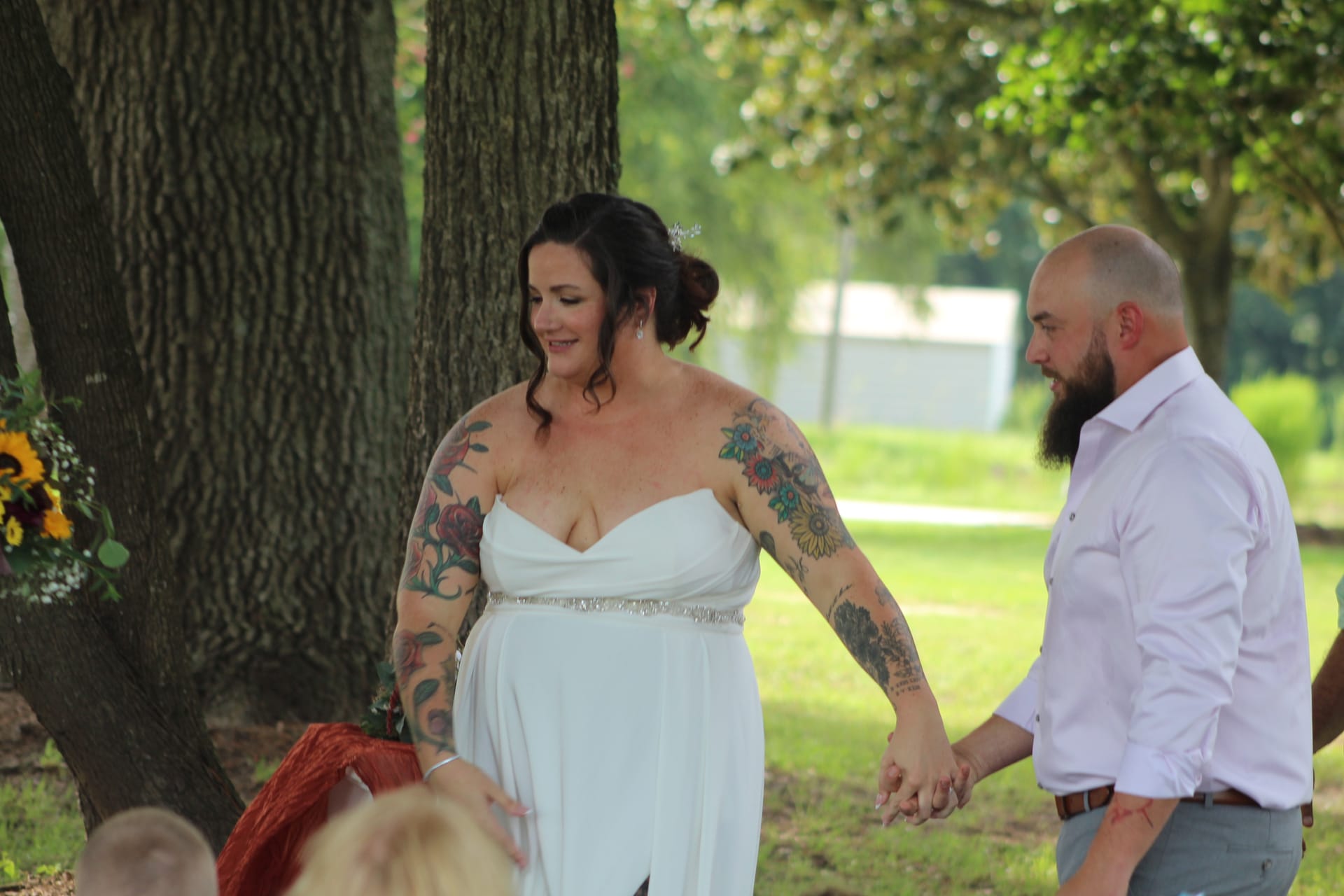 A tattooed bride in a white gown and her bearded groom hold hands and smile during an outdoor wedding ceremony beneath large oak trees at a historic estate venue.
