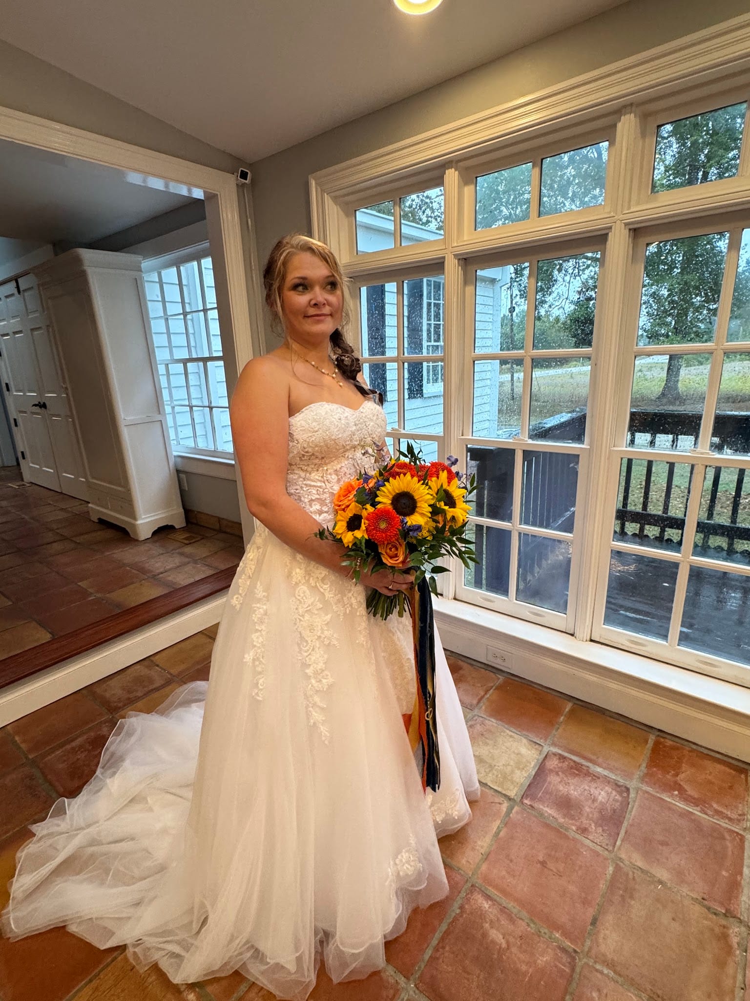 Bride in a strapless lace A-line gown holding a colorful sunflower and rose bouquet stands before large white-framed windows at Scarborough House historic estate wedding venue.