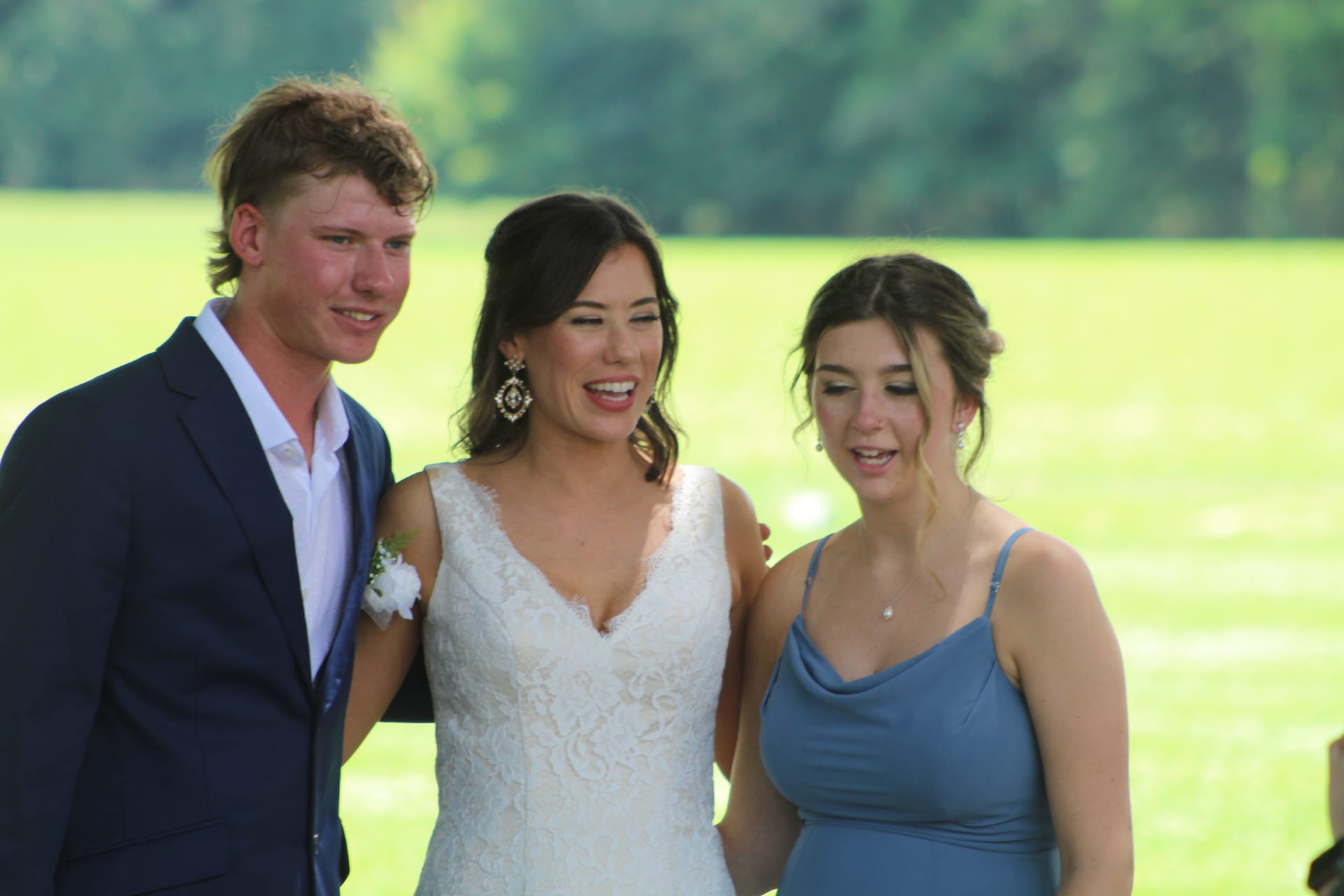 A laughing bride in a white lace gown shares a candid moment with a groomsman in a navy suit and a bridesmaid in a dusty blue dress on the sunlit ceremony lawn at Scarborough House.