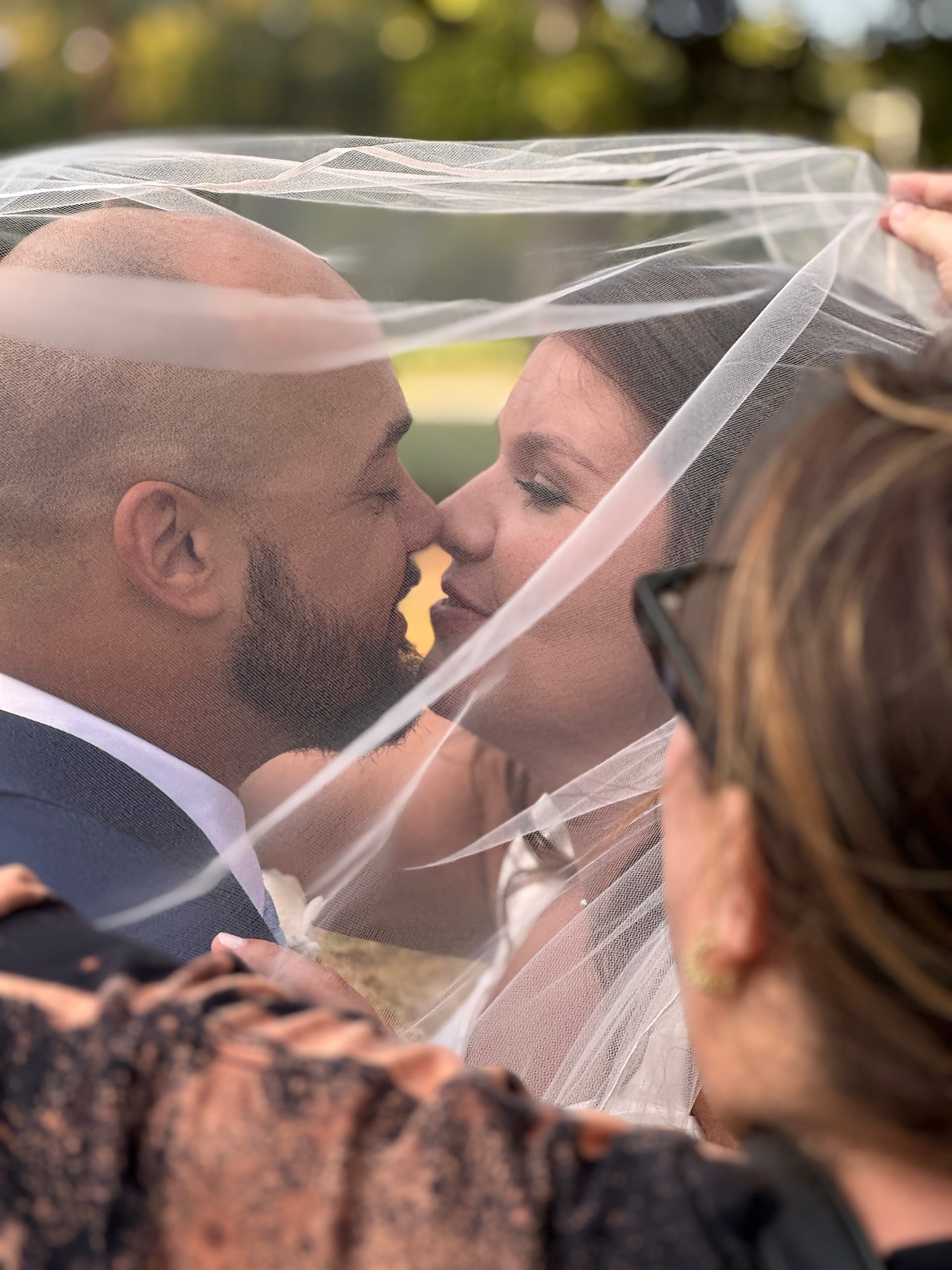 Bride and groom share a tender kiss beneath a flowing veil lifted by an attendant on the sunlit ceremony lawn at Scarborough House.