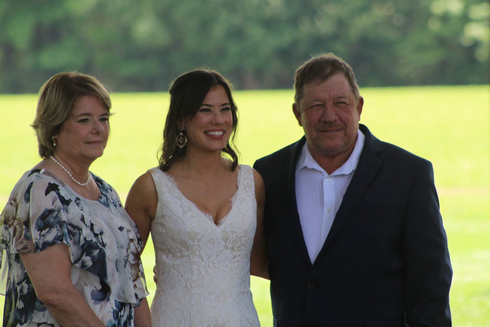 Smiling bride in a lace wedding gown poses outdoors with an older couple in front of a lush green lawn and trees at Scarborough House estate.