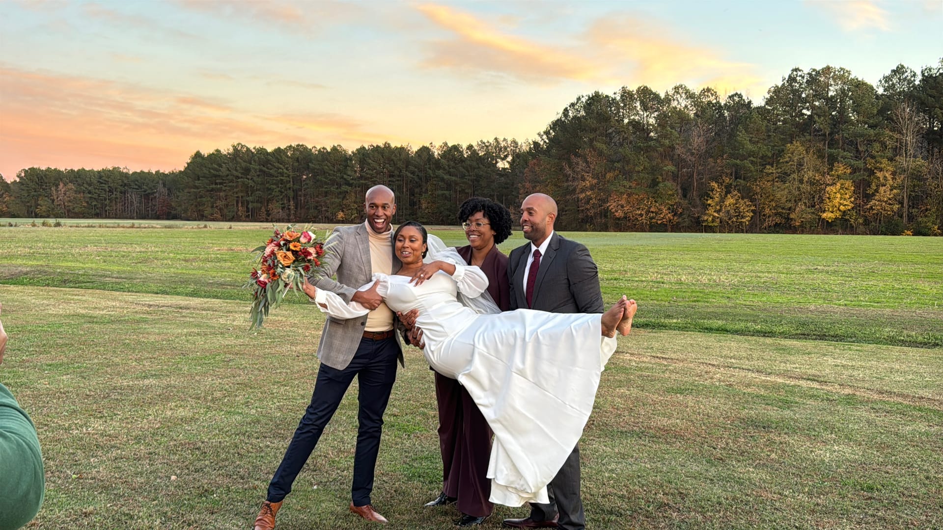 Bride in a white gown is playfully lifted by two groomsmen while a guest stands beside them smiling on a lush green field at sunset during an autumn wedding at Scarborough House.