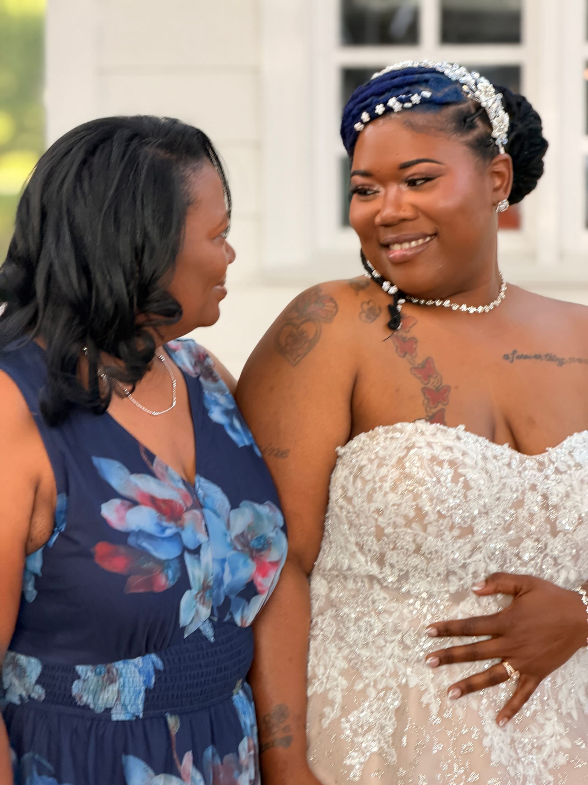 A smiling bride in a strapless beaded gown and jeweled headpiece shares a heartfelt moment with a guest in a navy floral dress outside the historic Scarborough House estate.