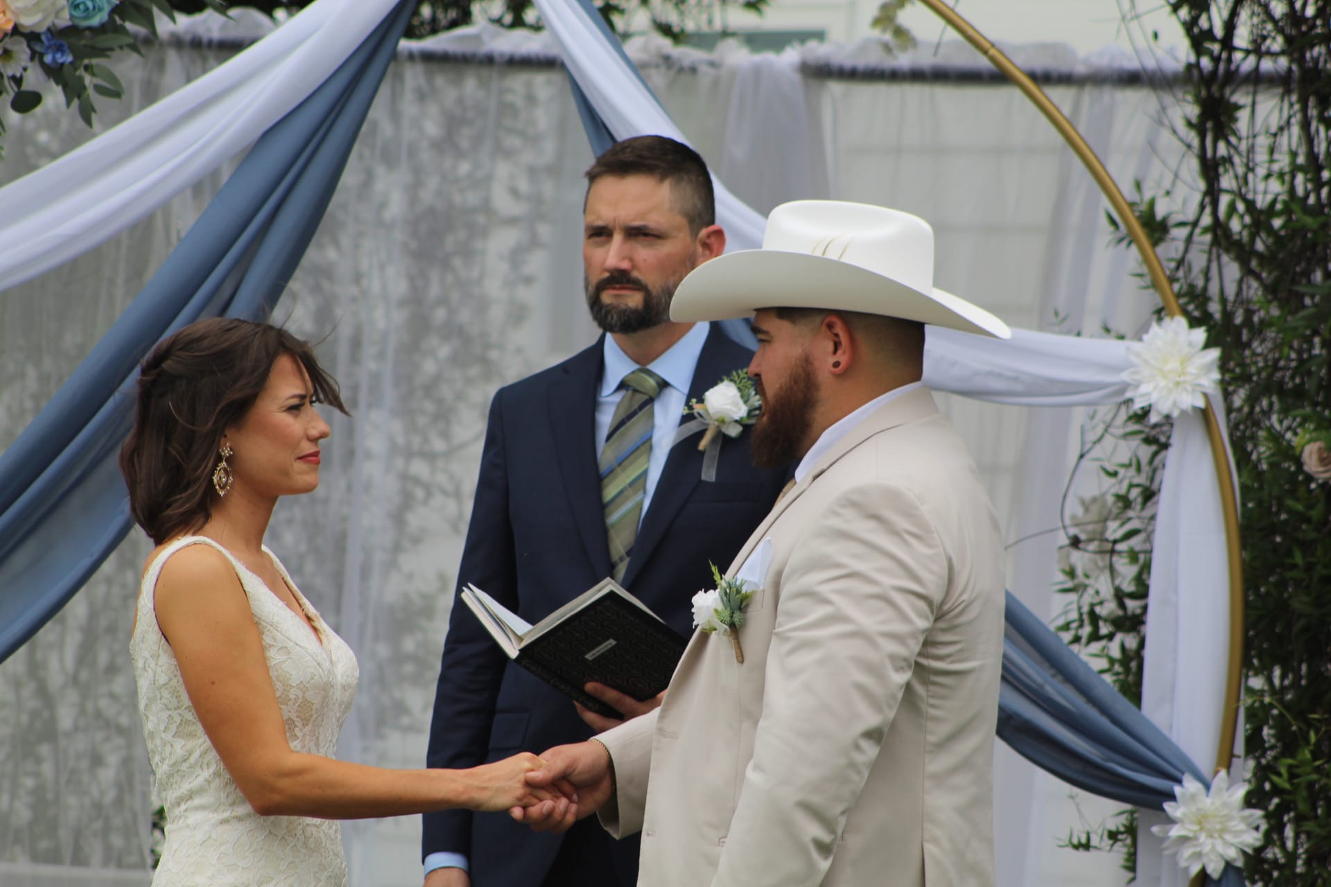 Bride and groom hold hands during vow exchange beneath a gold circular arch draped in blue fabric and white florals while the officiant reads from a book at Scarborough House wedding venue.