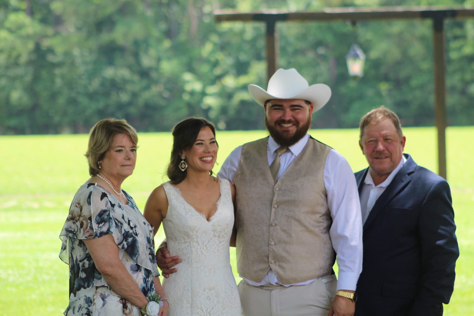 Bride in lace gown and groom in tan vest and white cowboy hat pose with two family members on the ceremony lawn at Scarborough House wedding venue.