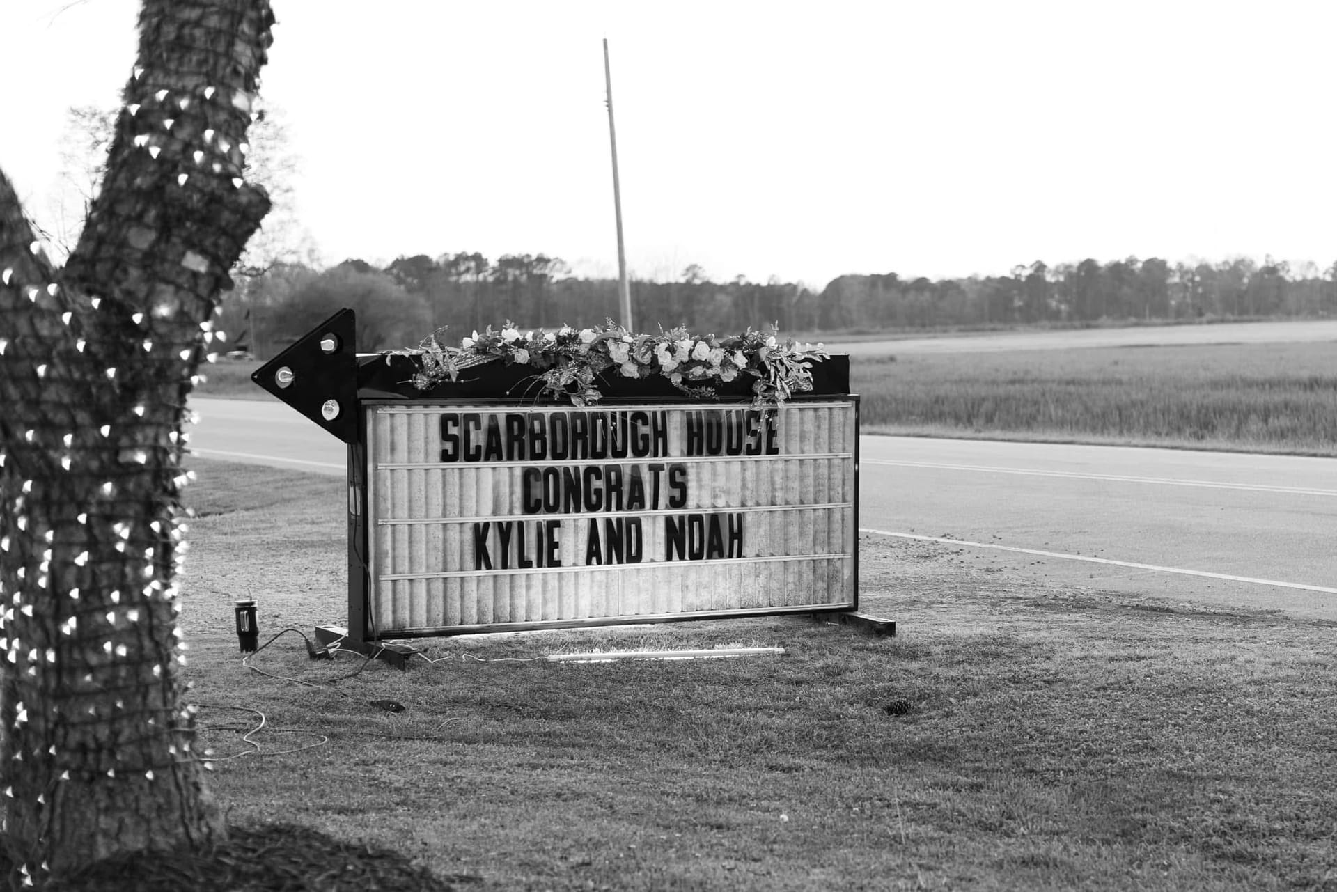 Black and white photo of the Scarborough House illuminated marquee sign decorated with floral greenery congratulating wedding couple Kylie and Noah, set along a rural roadside with farmland fields in the background.