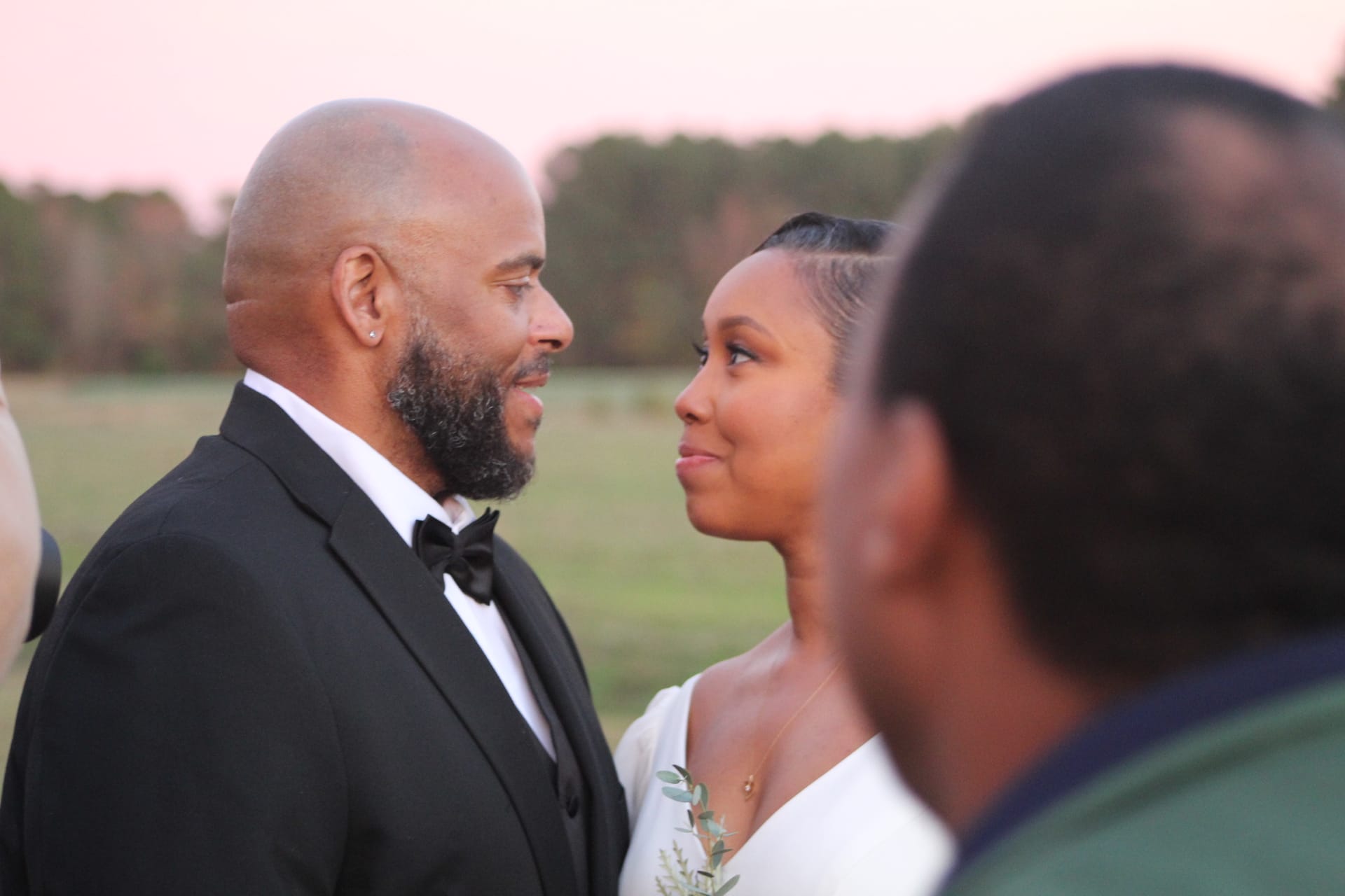 Bride and groom share a loving glance during their outdoor wedding ceremony at Scarborough House, bathed in soft pink sunset light with open farmland fields in the background.