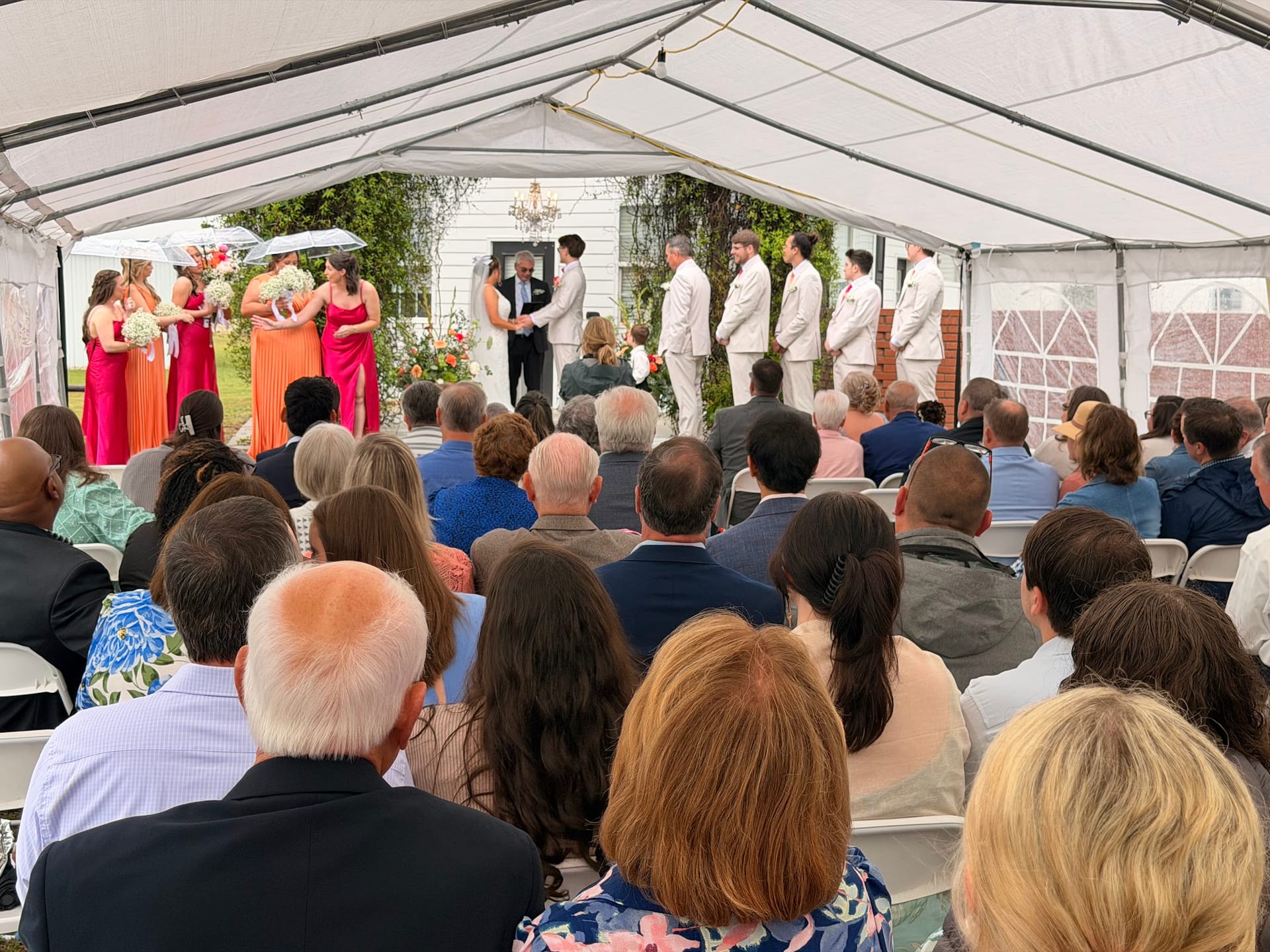 Wedding ceremony under a white marquee tent at Scarborough House with guests seated facing the altar where the couple exchanges vows surrounded by colorful bridesmaids in orange and pink dresses and groomsmen in tan suits.
